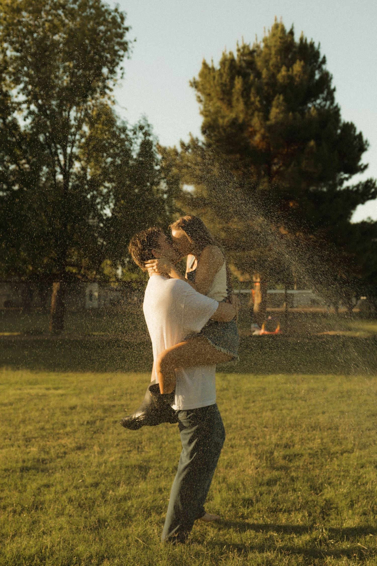 Romantic couple kissing and embracing in a backyard sprinkler shower during warm golden hour sunlight.