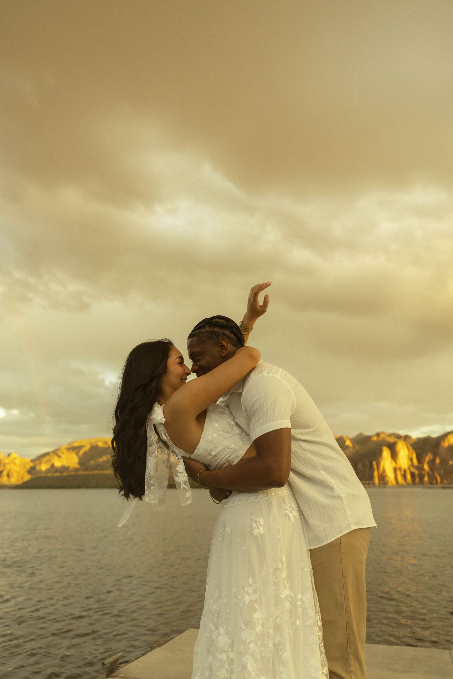 Interracial couple sharing a kiss on a dock at golden sunset with mountains and water behind them.