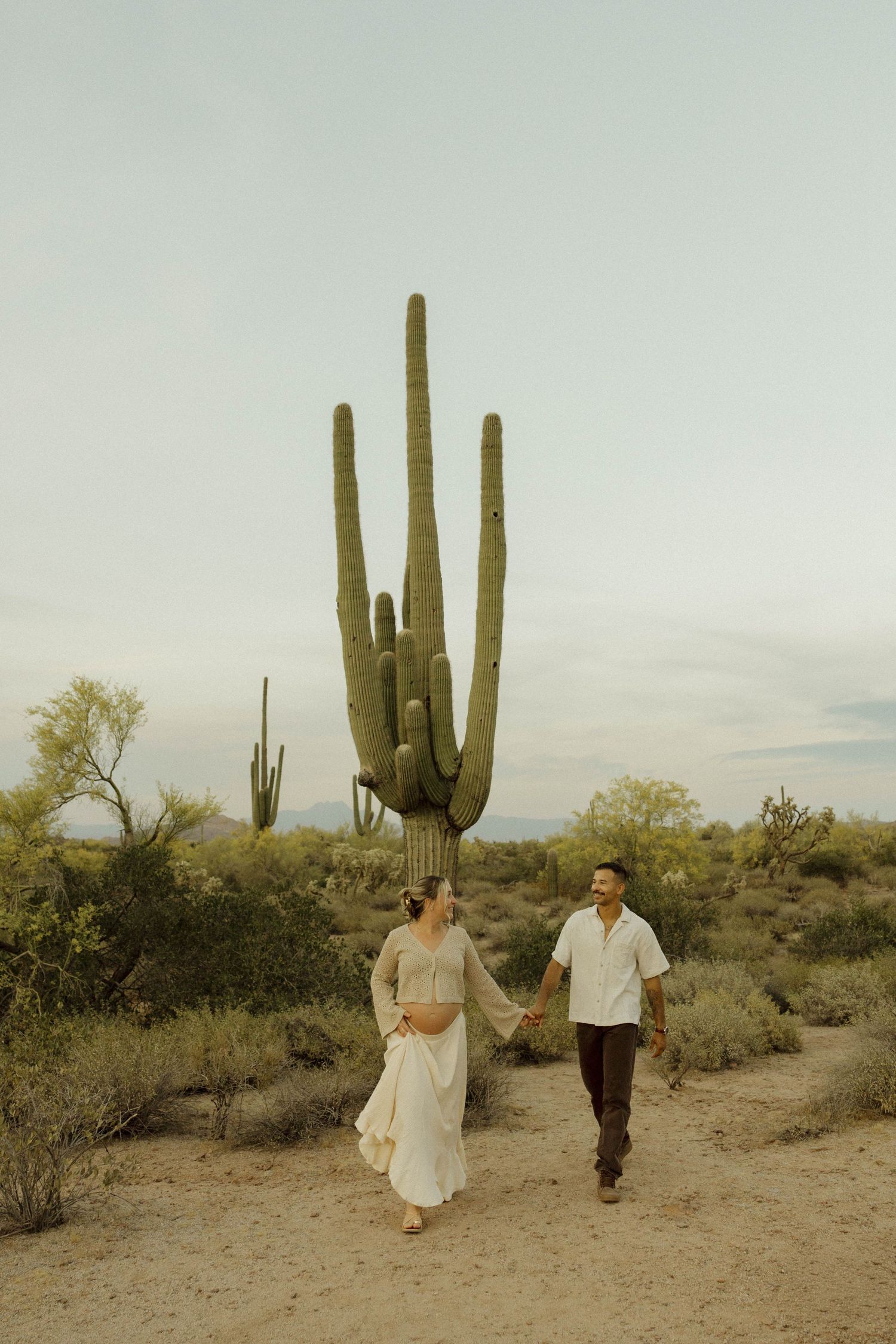 Couple holding hands walking in Arizona desert with tall saguaro cactus backdrop during golden hour maternity photoshoot.