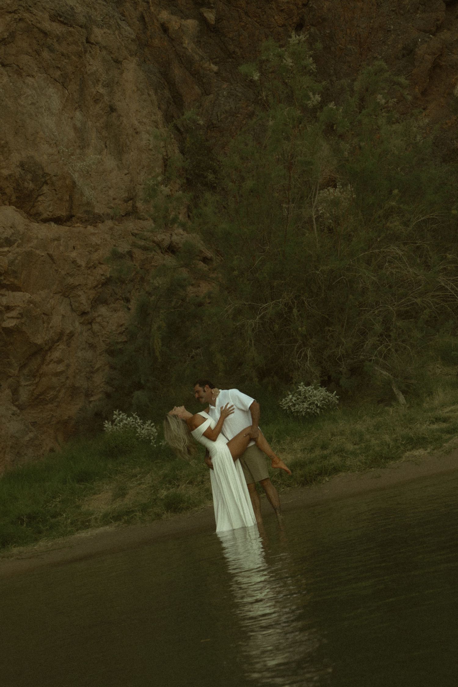 Couple sharing a romantic embrace in shallow water near rocky cliffs and lush greenery, wearing white outfits.