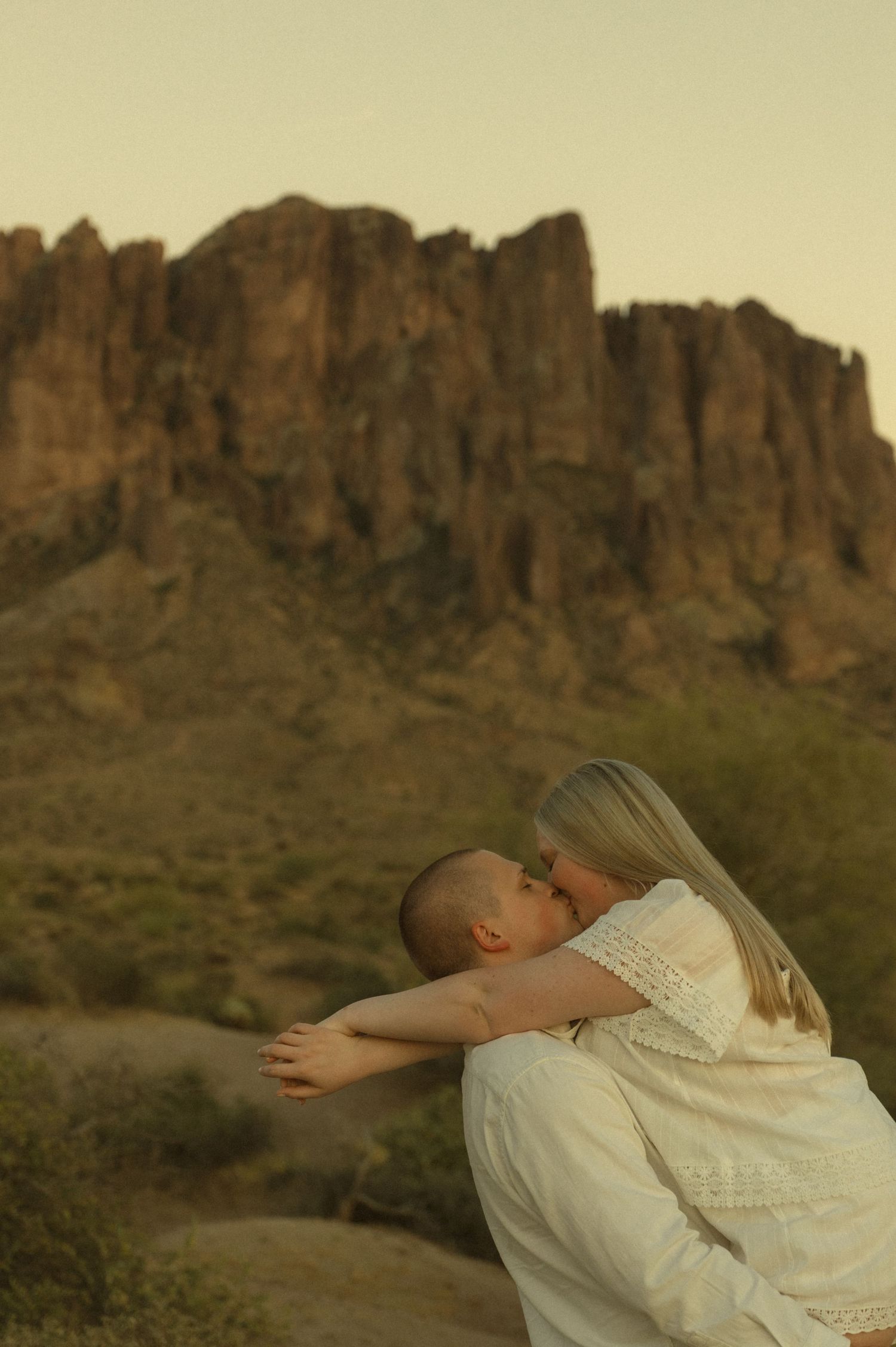 Couple sharing a romantic kiss with stunning desert mountain landscape in the background at golden hour.
