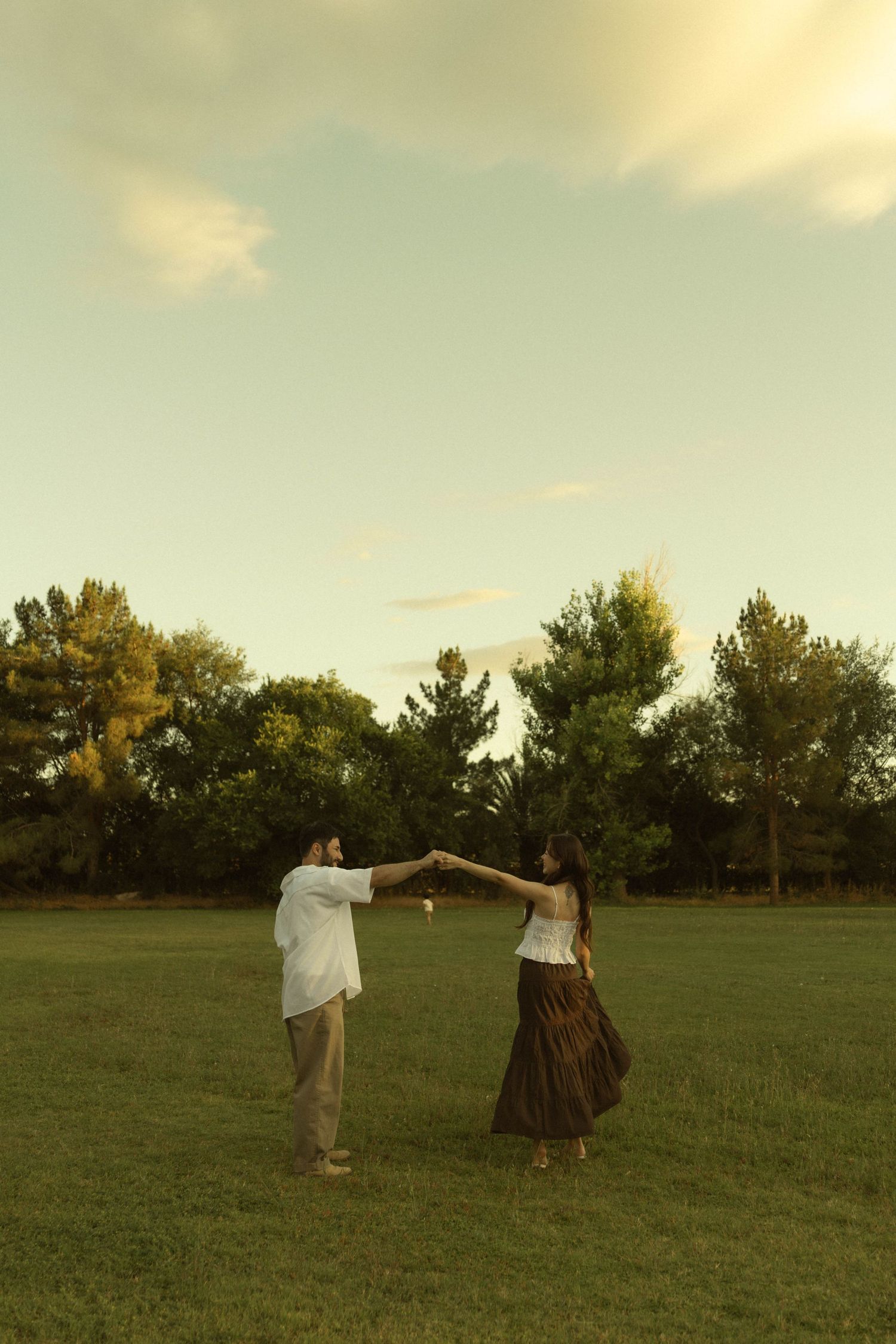Couple dancing together in a green field at sunset, holding hands with trees in the background.