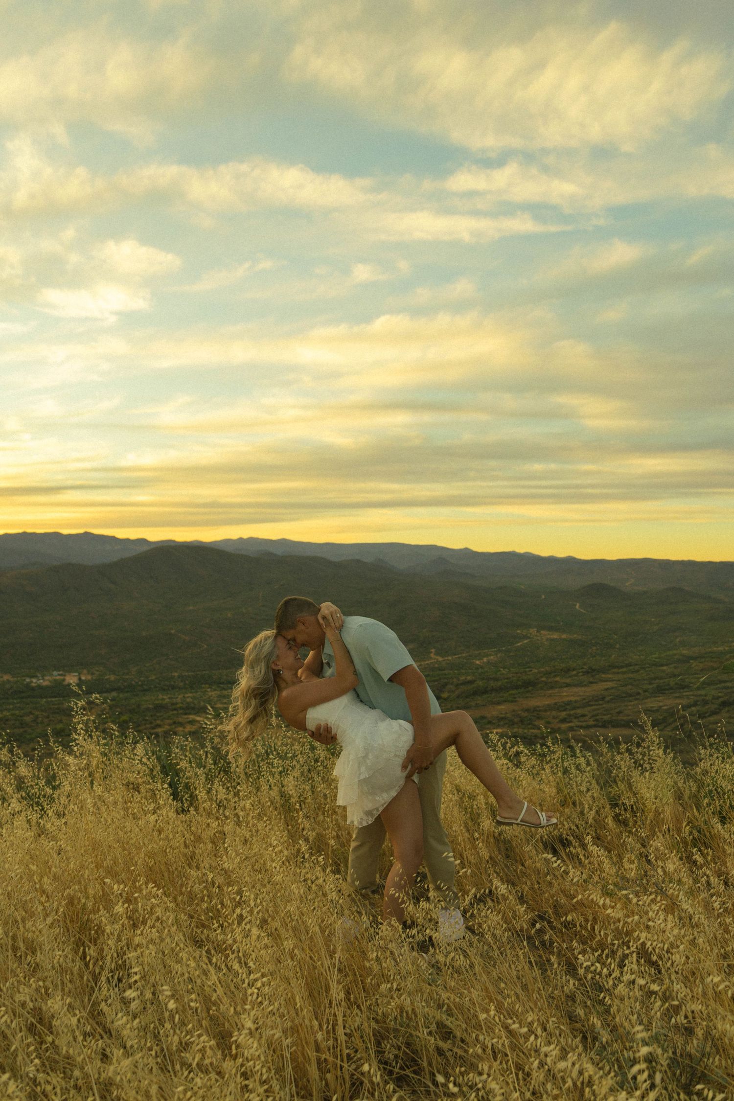 Couple sharing a romantic dip kiss in golden grass field at sunset with mountain views.