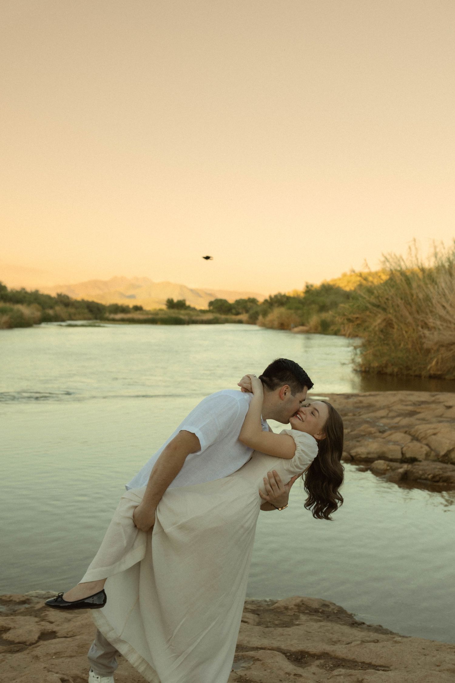 Couple sharing a romantic dip kiss by a serene river at golden sunset.