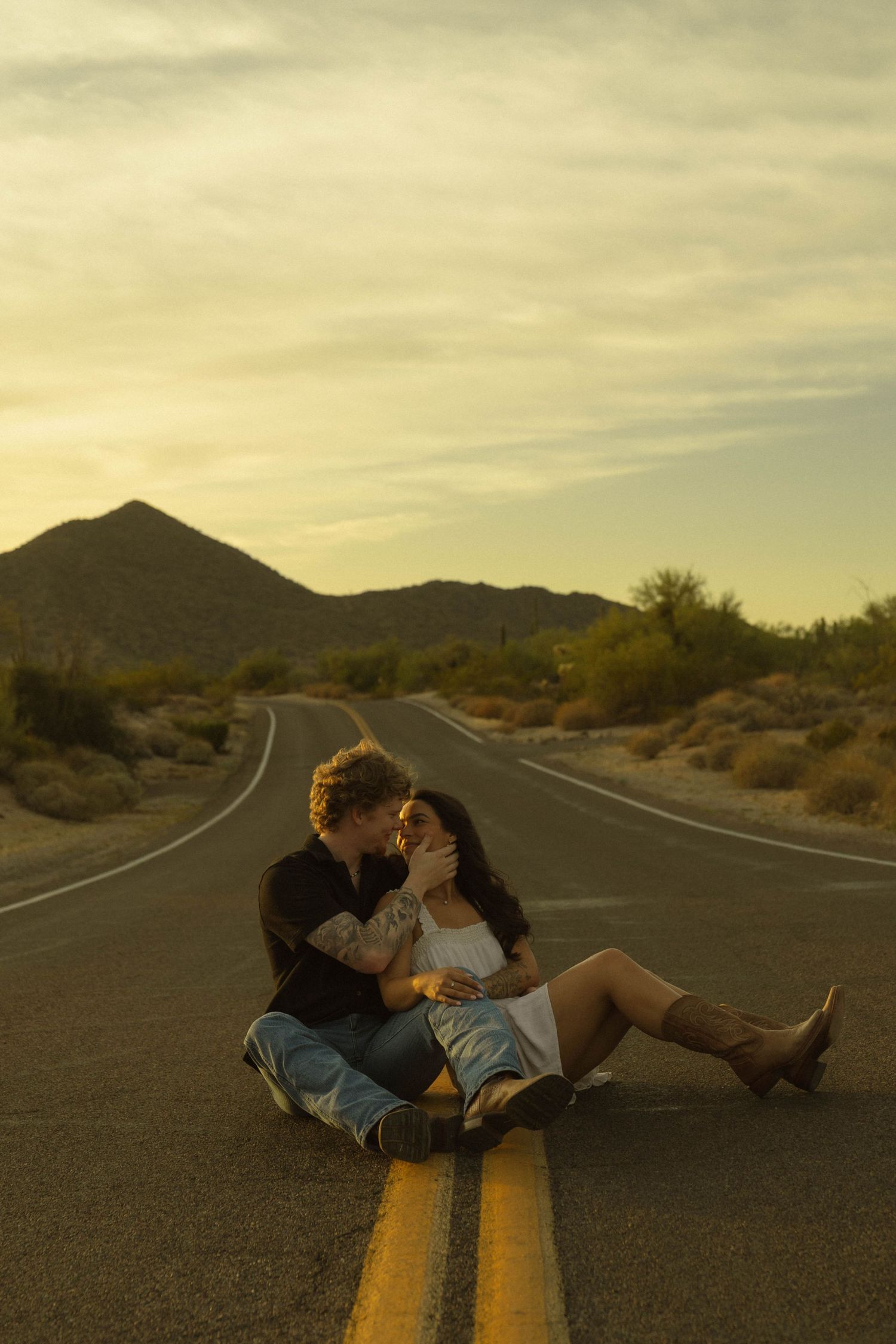 Couple sitting on desert road centerline at sunset with mountains in background during engagement shoot.
