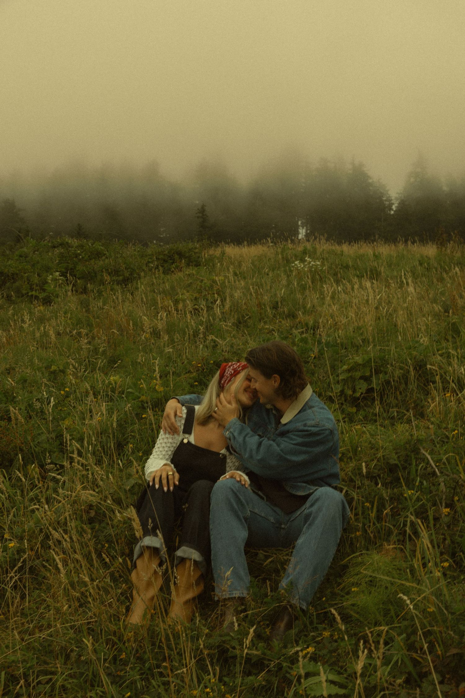 Couple sitting intimately in a misty green meadow, embracing romantically with foggy trees in background.