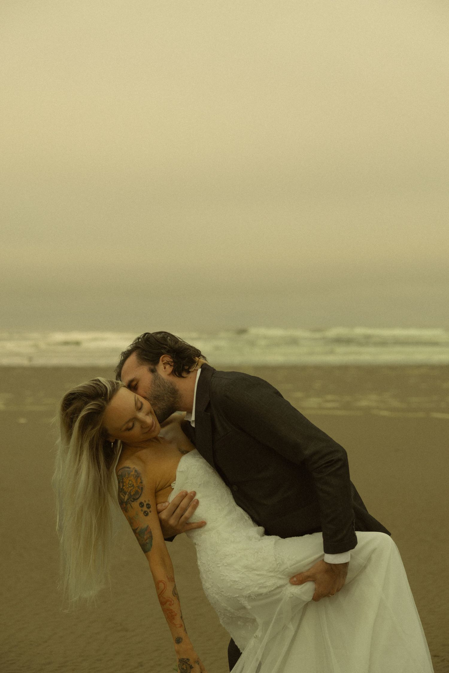 Groom dips tattooed bride for a romantic kiss on a misty Oregon beach in wedding attire.