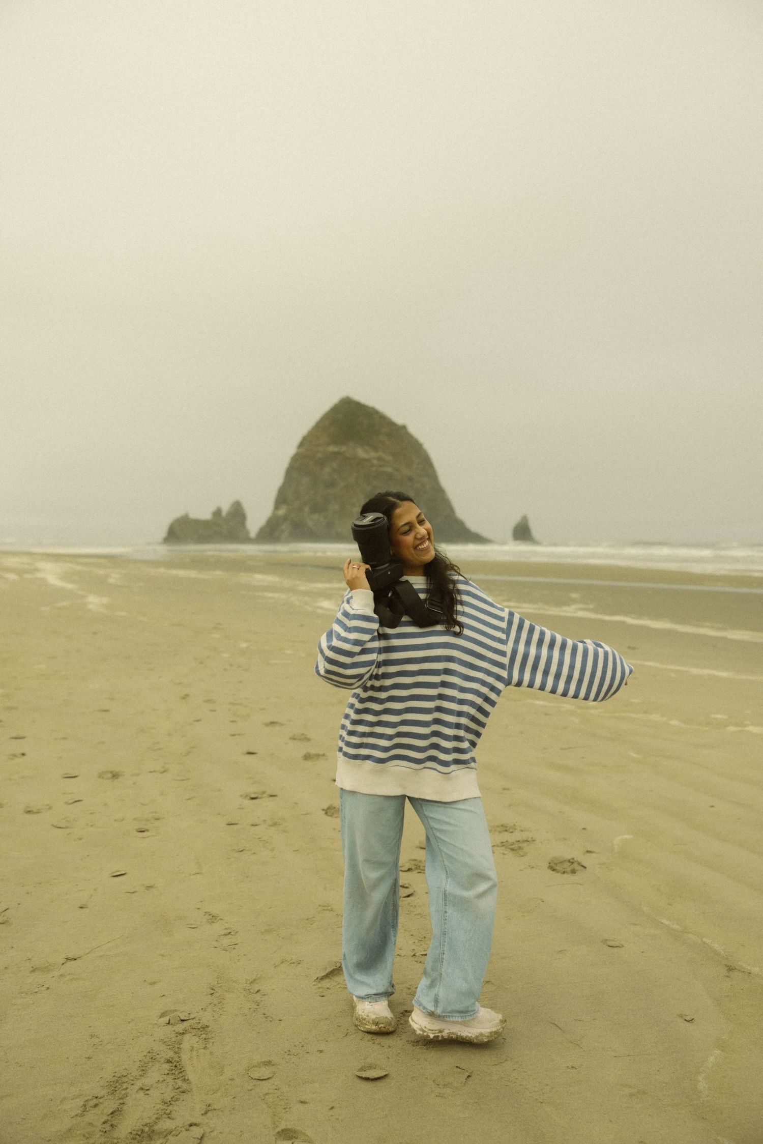 Woman in striped sweater and jeans posing on a foggy beach with Haystack Rock in the background.