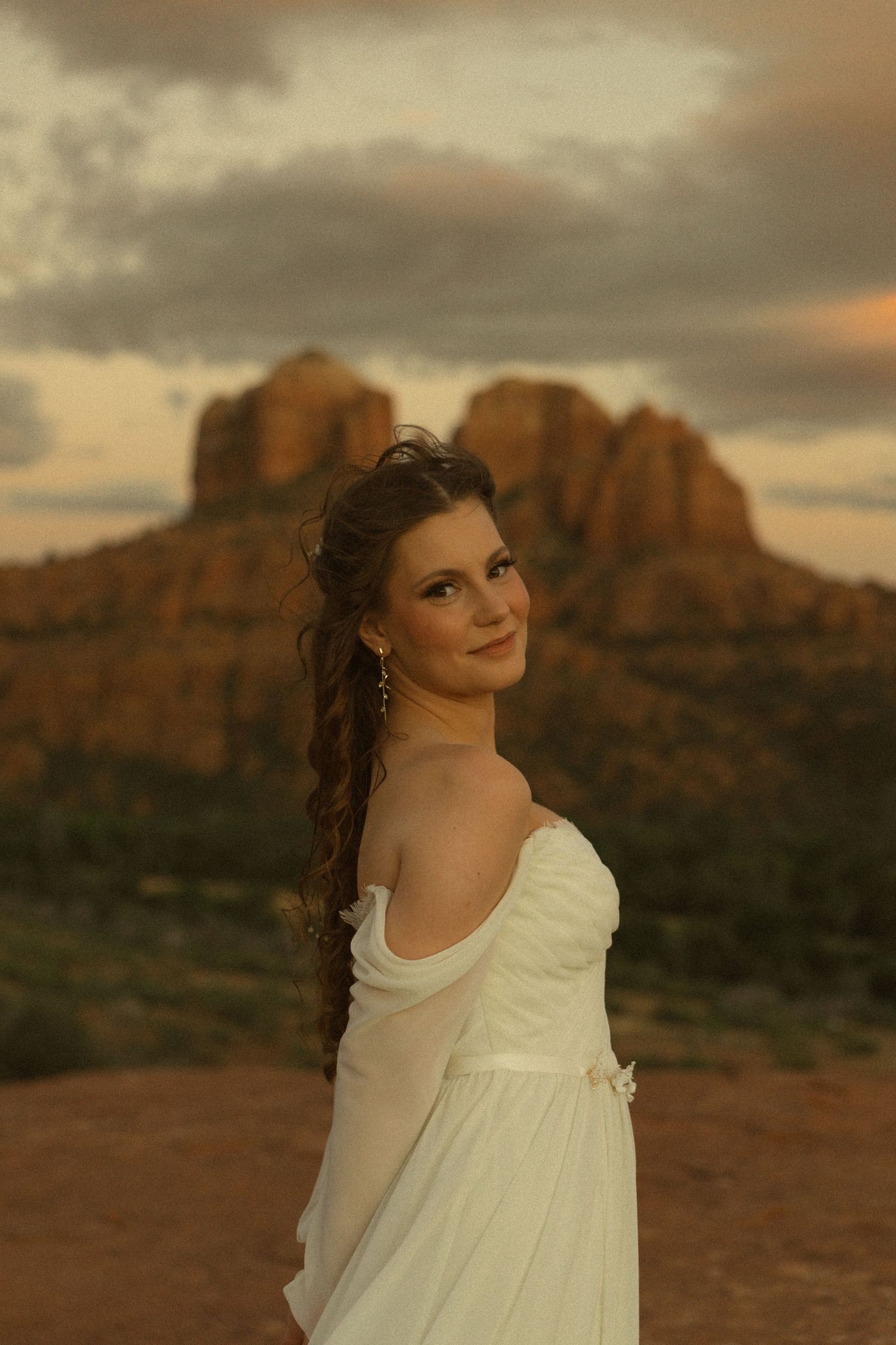 Young woman in white dress poses against Sedona red rocks at golden sunset, hair in long braid, smiling over shoulder.