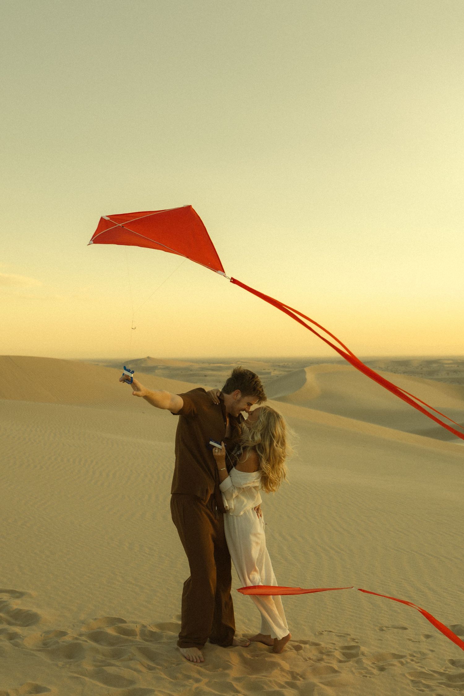 Couple kissing while flying a red kite on sandy desert dunes at golden sunset.