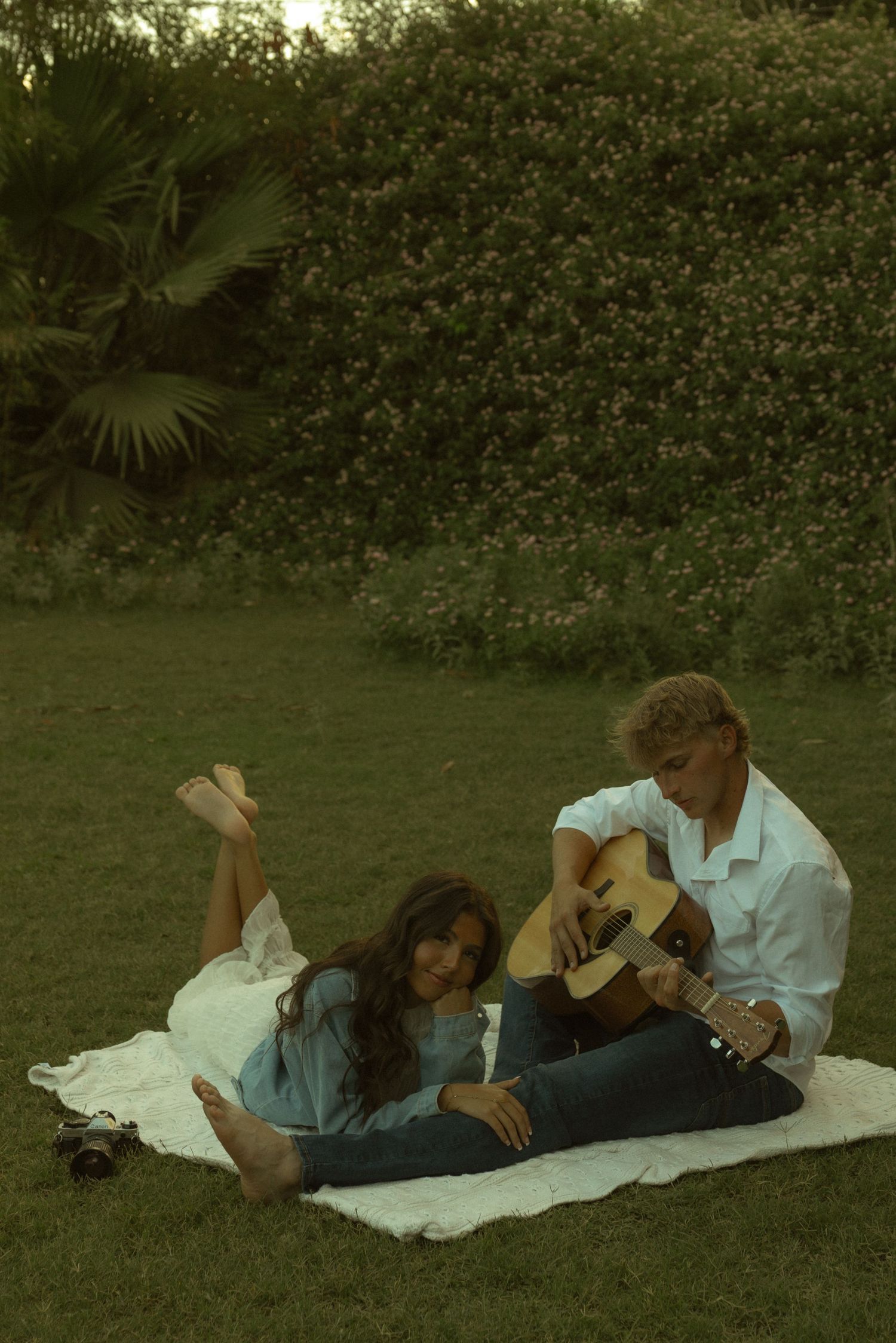 Couple relaxing on white blanket in lush garden, man playing acoustic guitar to woman lying beside him.