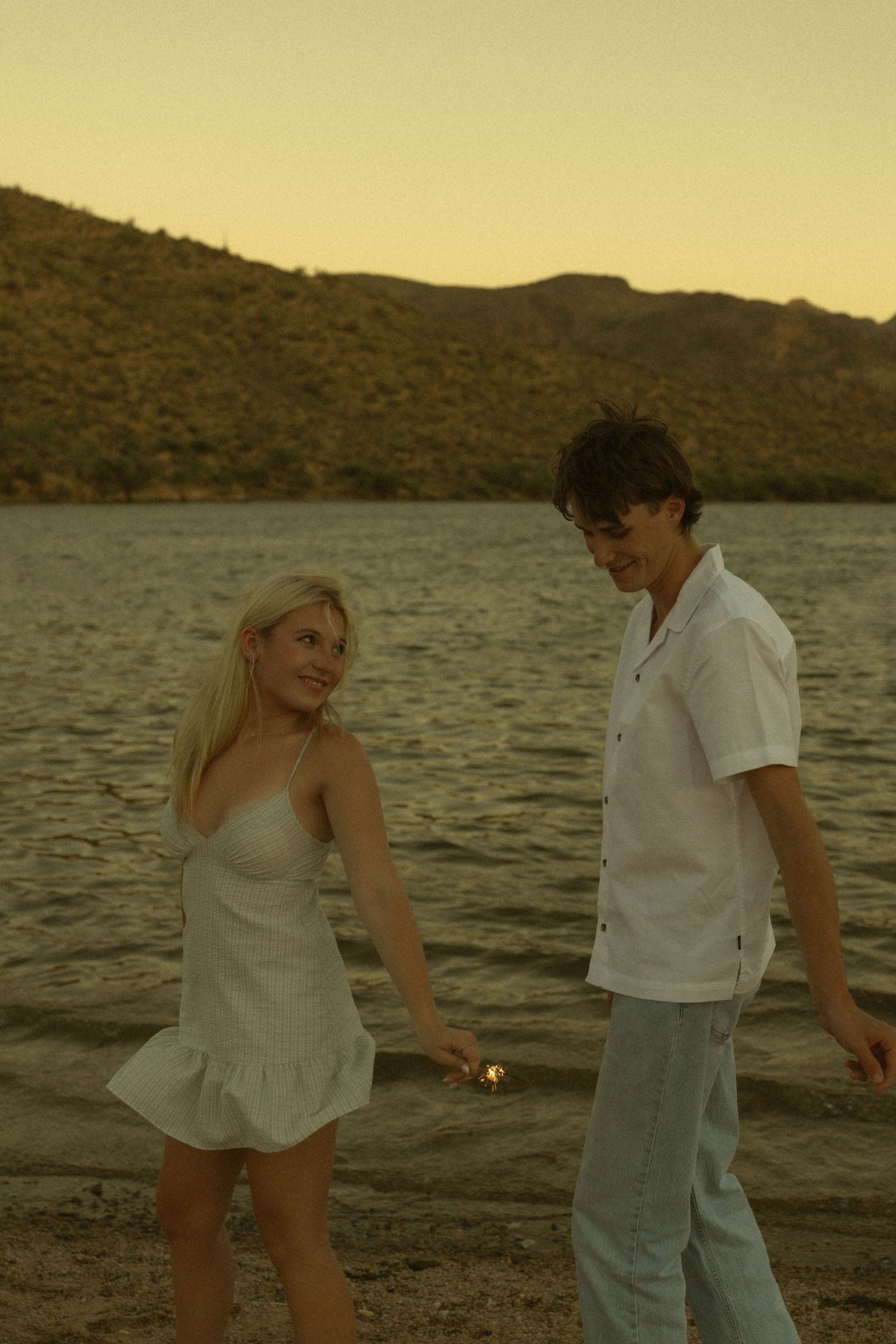 Young couple holding hands by a lake at sunset, woman in white dress, man in white shirt, mountains in background.