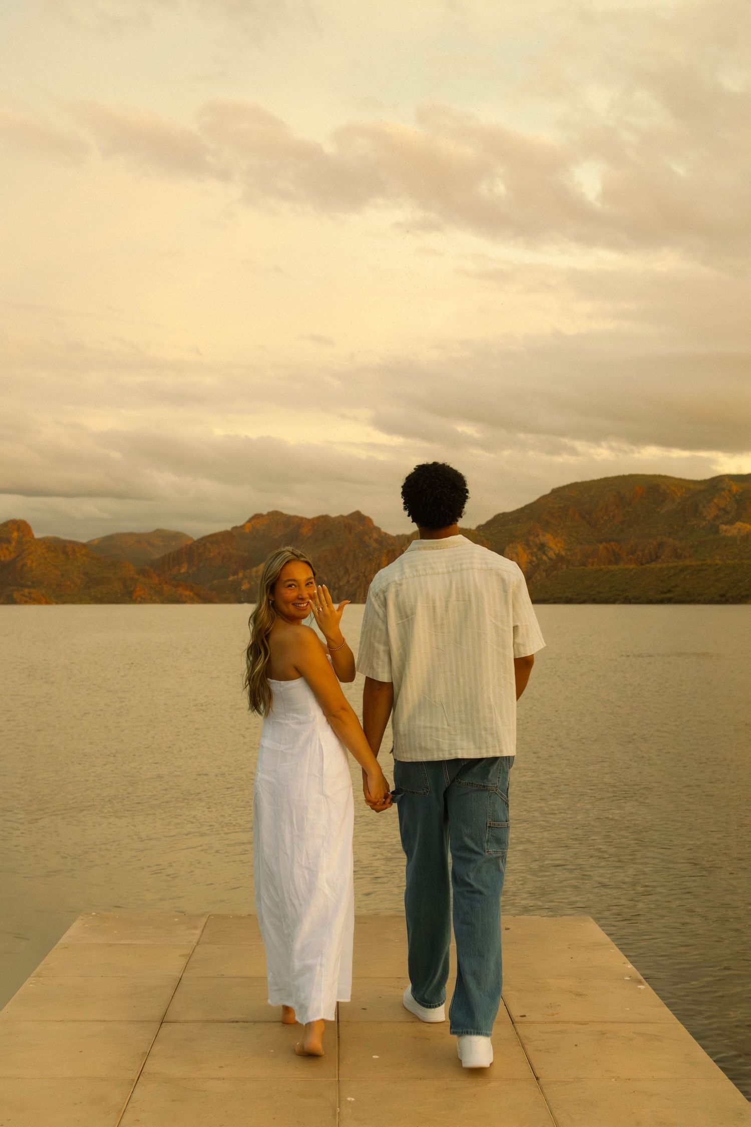 Couple walking away on a dock at sunset with mountains and water in the background.