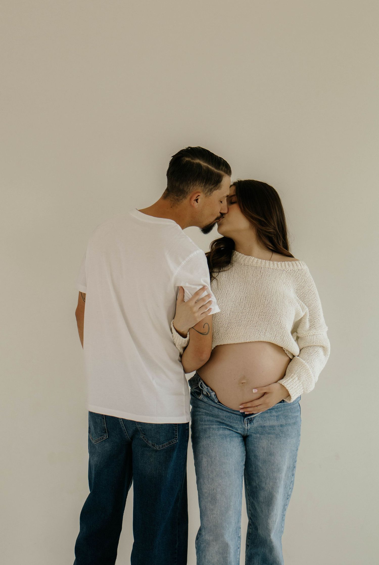 Romantic maternity photo of couple in white and denim sharing tender moment against beige background.