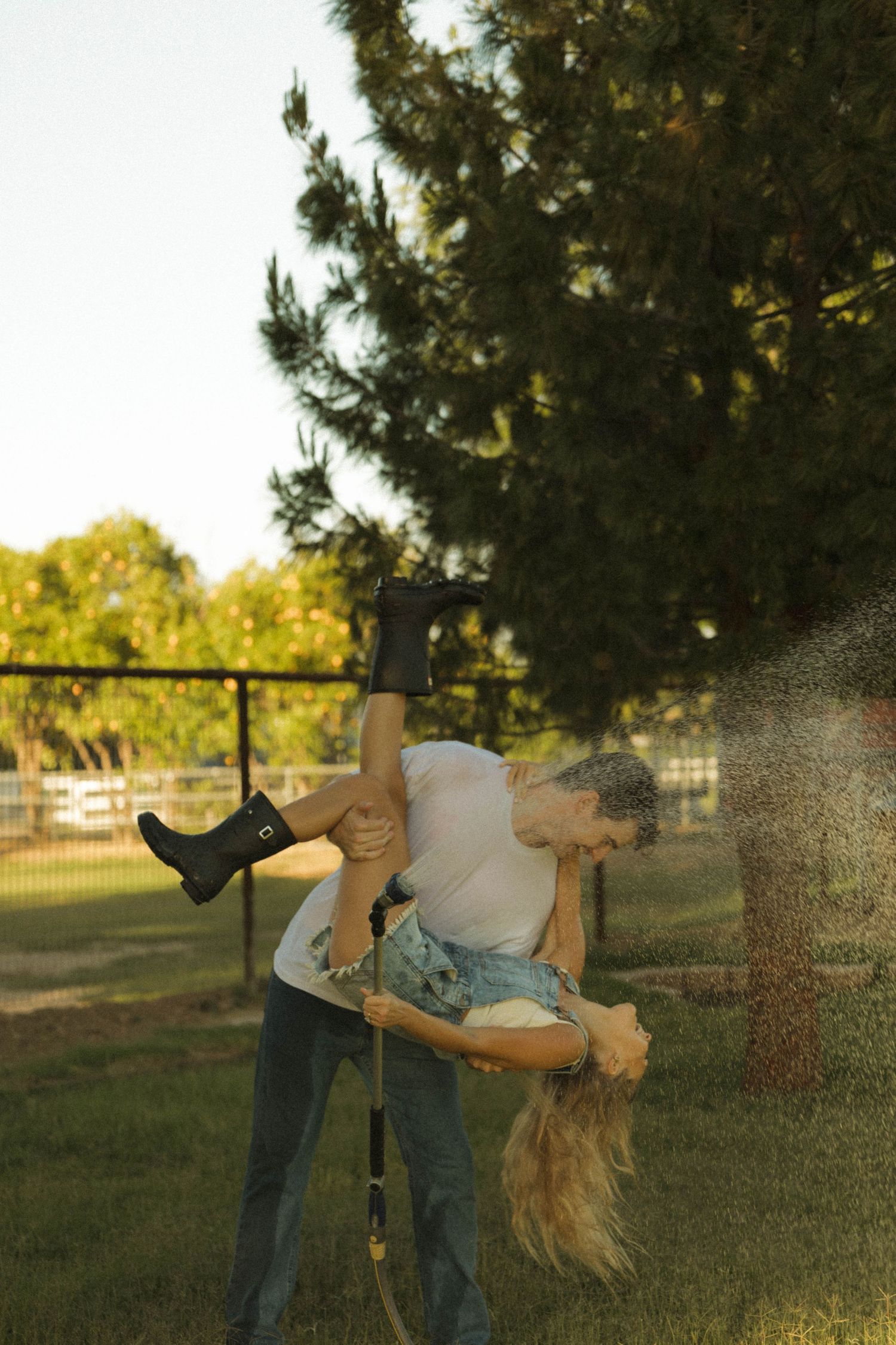 Couple playfully dancing with a garden hose in a sunny backyard near a tall pine tree.