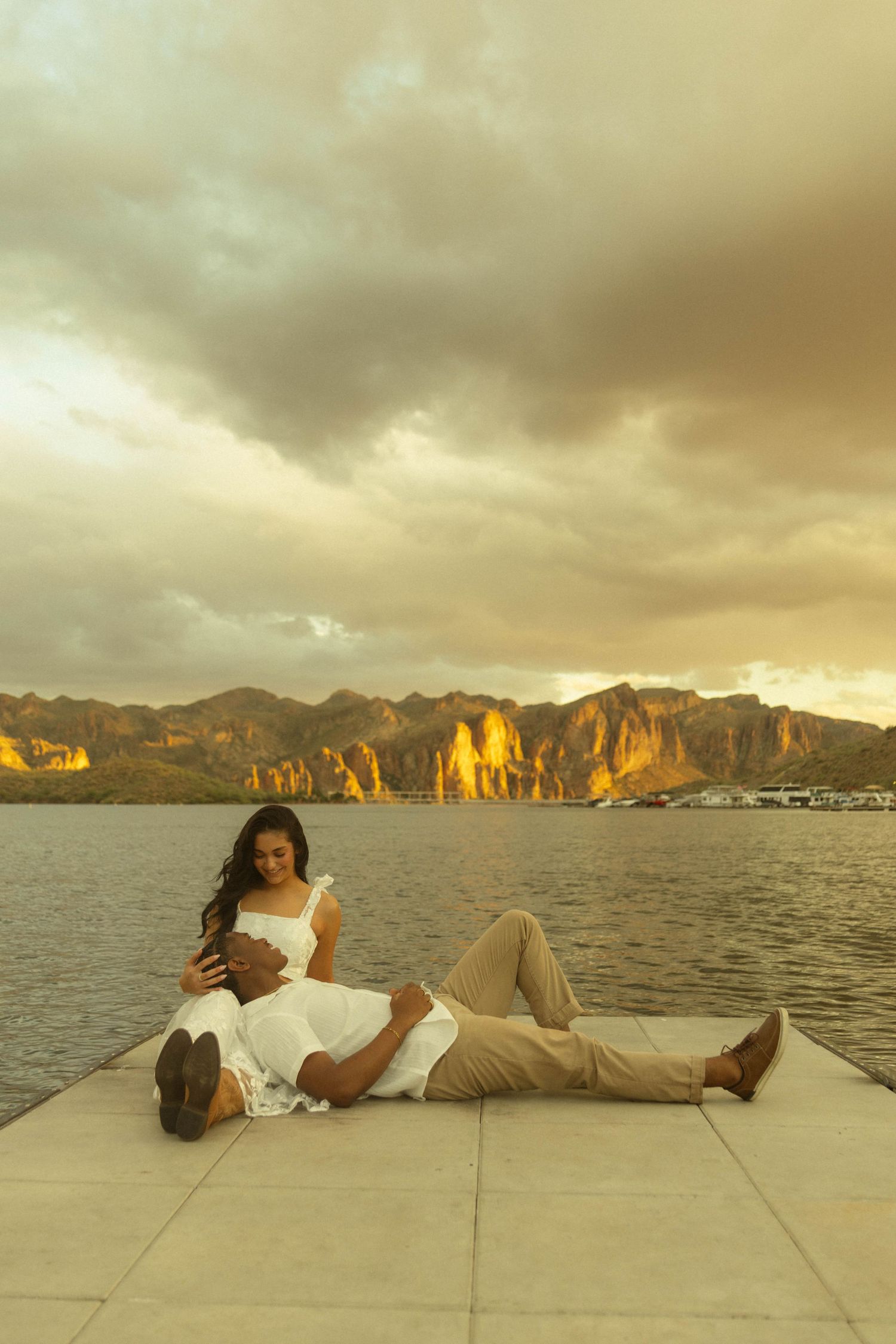 Couple relaxing on a waterfront dock at golden sunset with dramatic rocky mountains and cloudy sky backdrop.