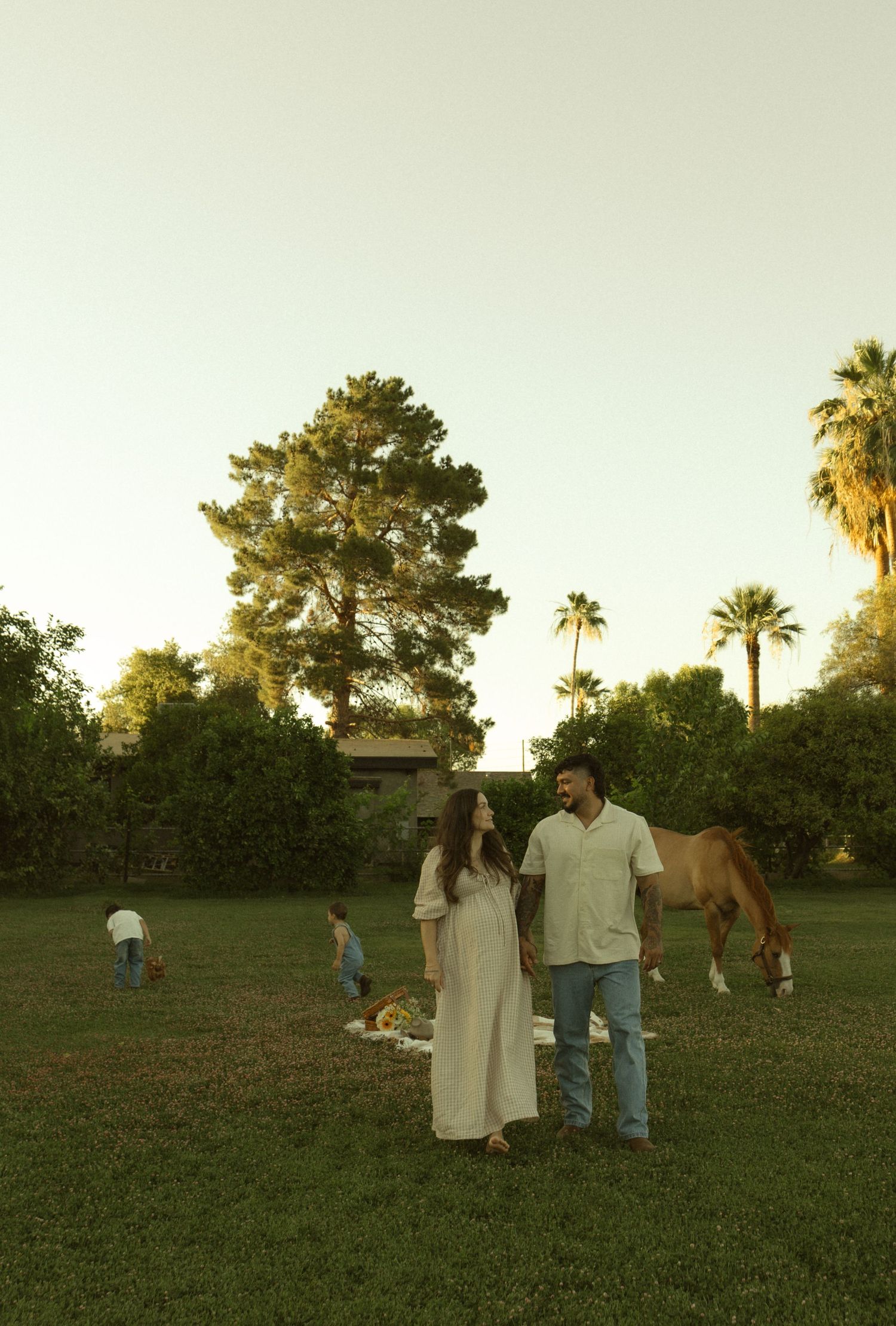 Couple standing on green lawn at golden hour with horse, children, and palm trees in background.