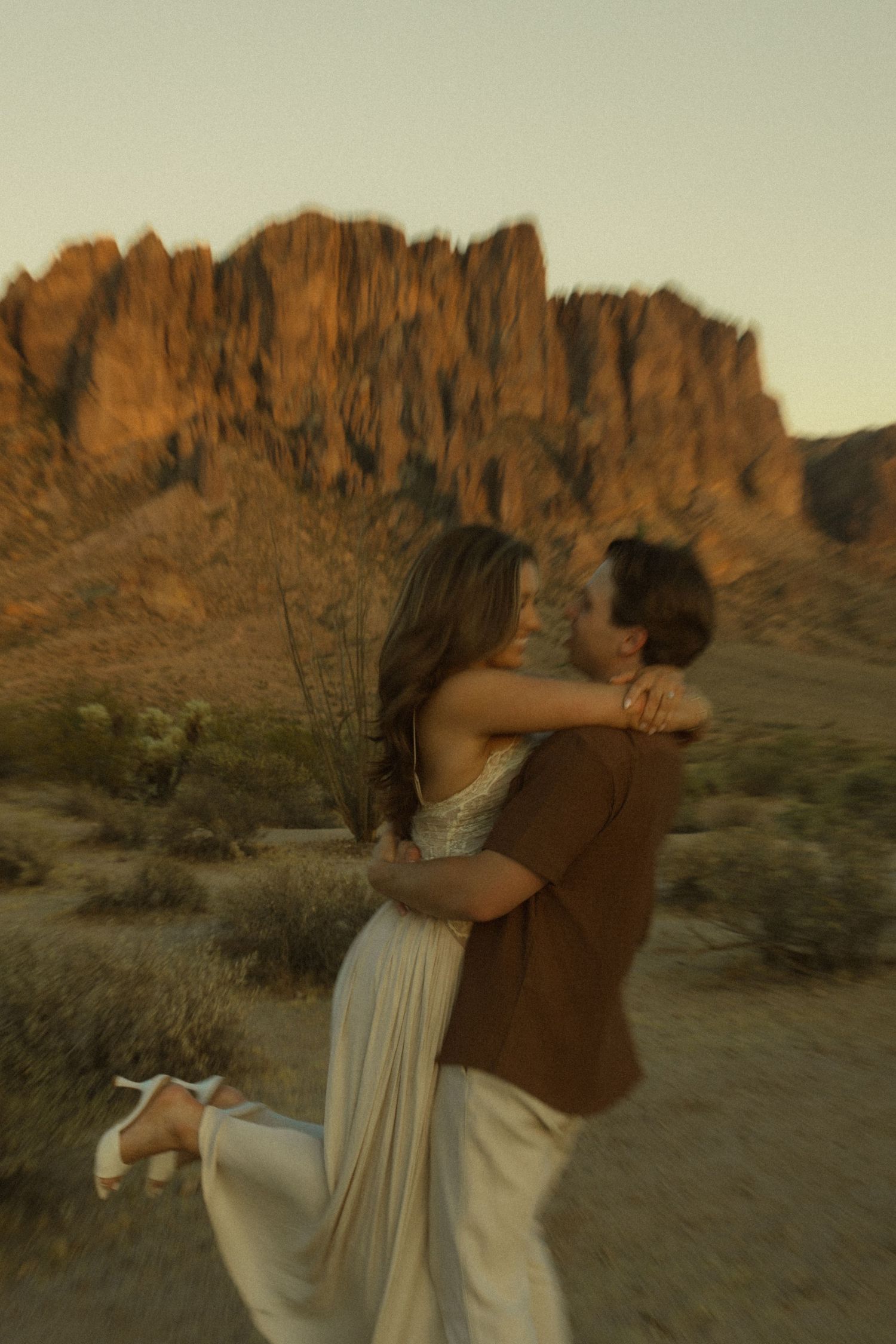 Couple embracing at golden hour in the desert with dramatic red rock mountains in the background.