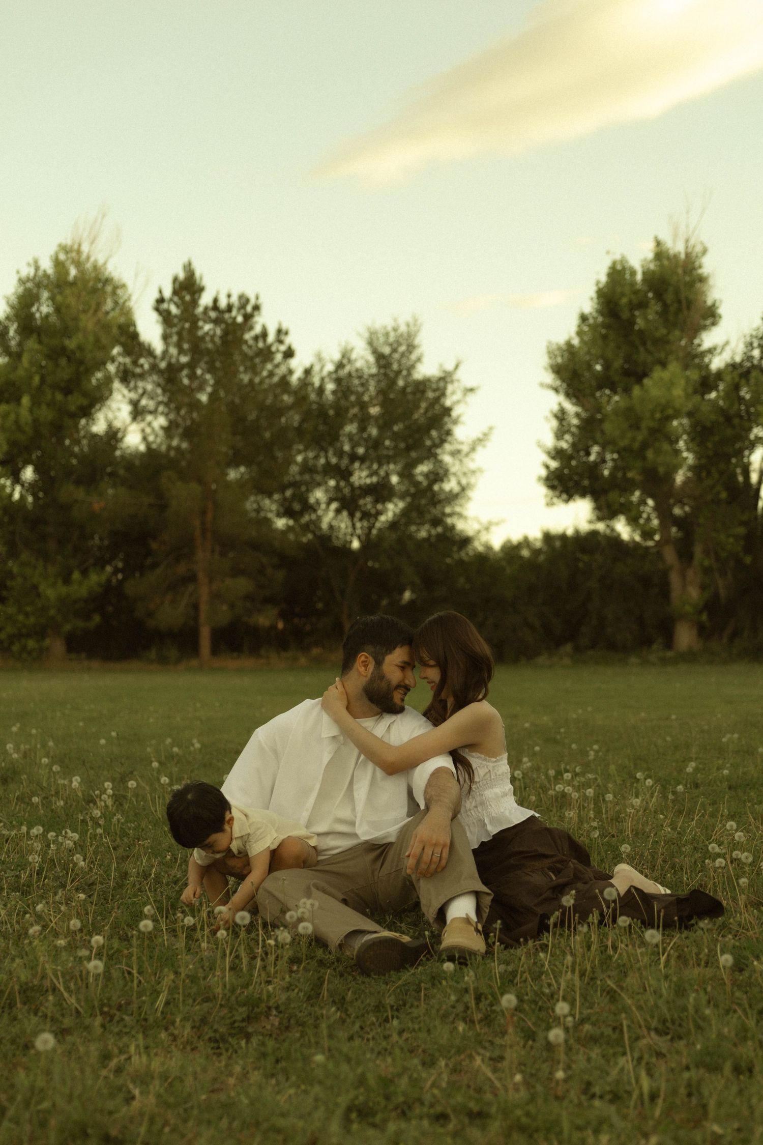 A couple sits romantically on a grass field at sunset, while a child plays nearby, surrounded by lush green trees.