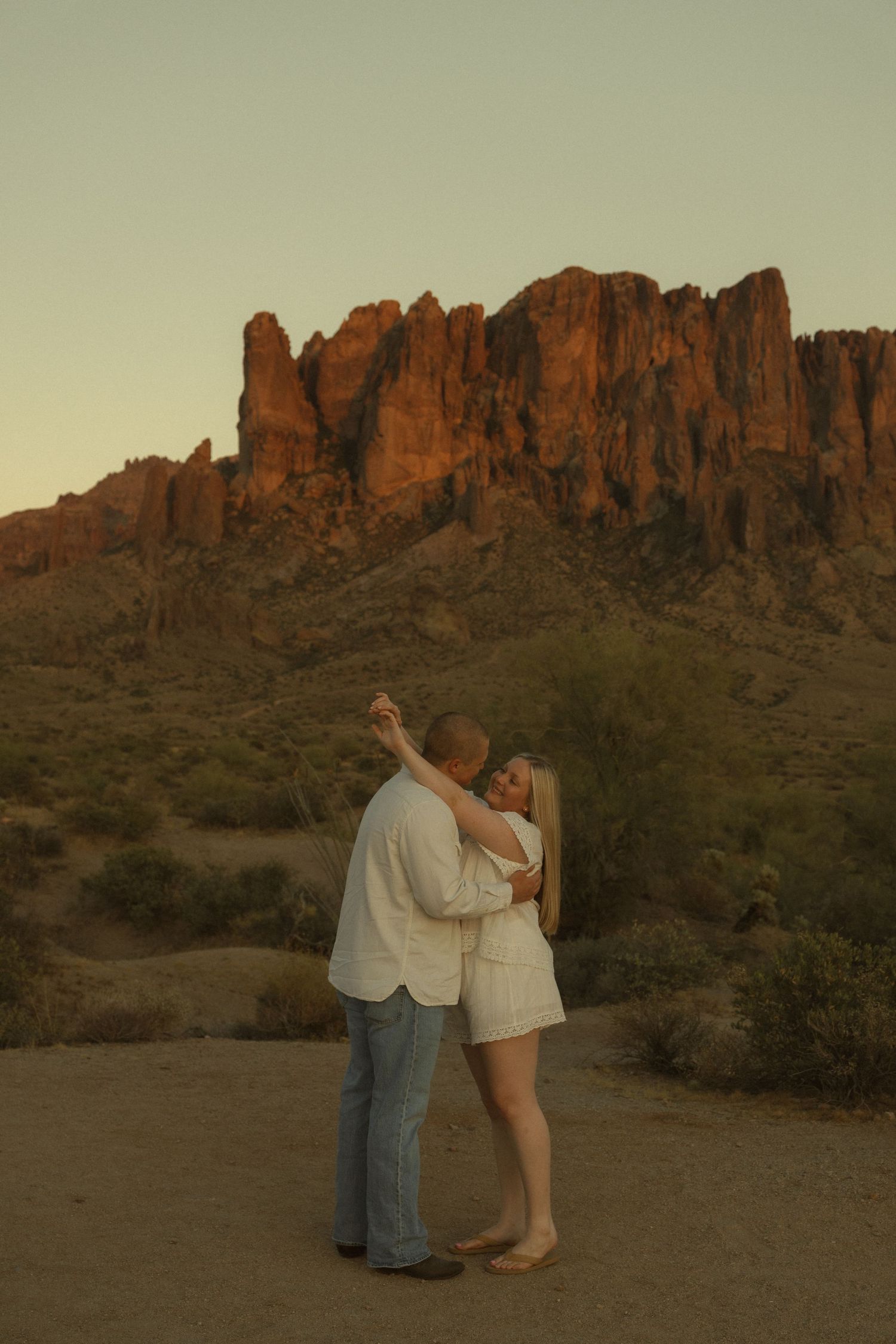 Couple embracing at sunset in Arizona desert with Superstition Mountains glowing red-orange in the background.
