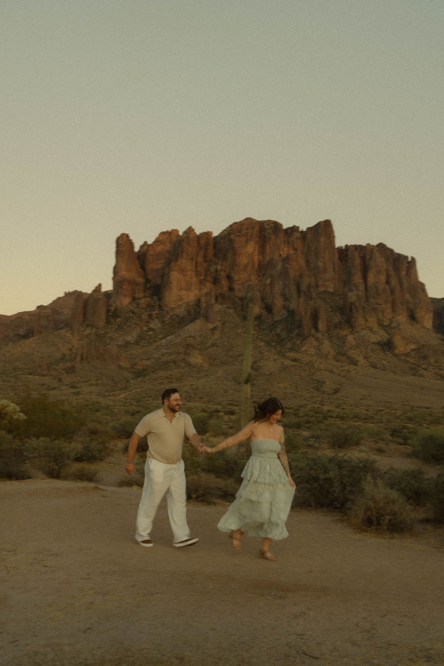 Couple holding hands walking in desert with Superstition Mountains at sunset.
