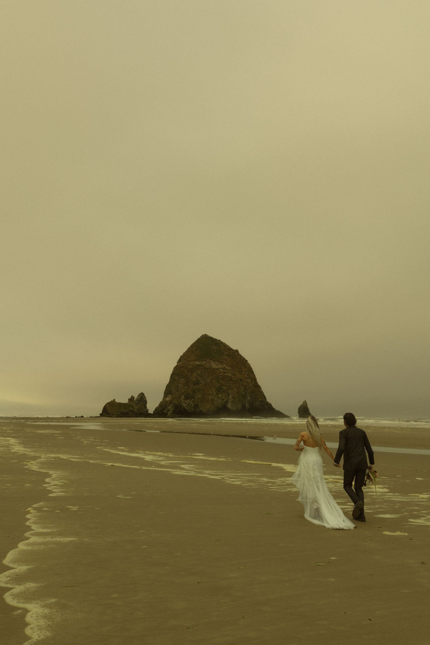 Bride and groom walking hand in hand along Cannon Beach shoreline toward Haystack Rock.