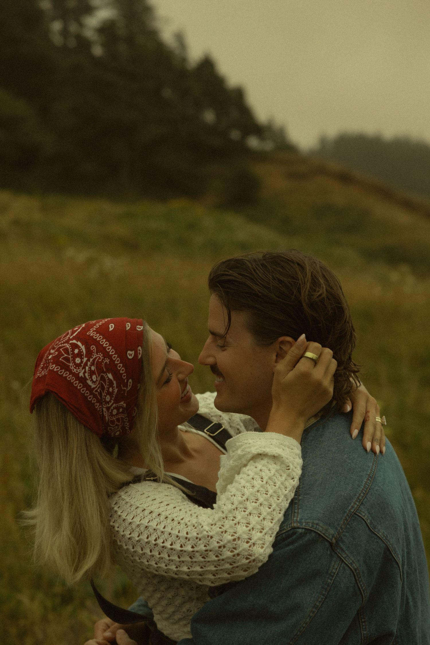 A couple embraces and smiles in a misty green field, the woman wearing a red bandana and white sweater.