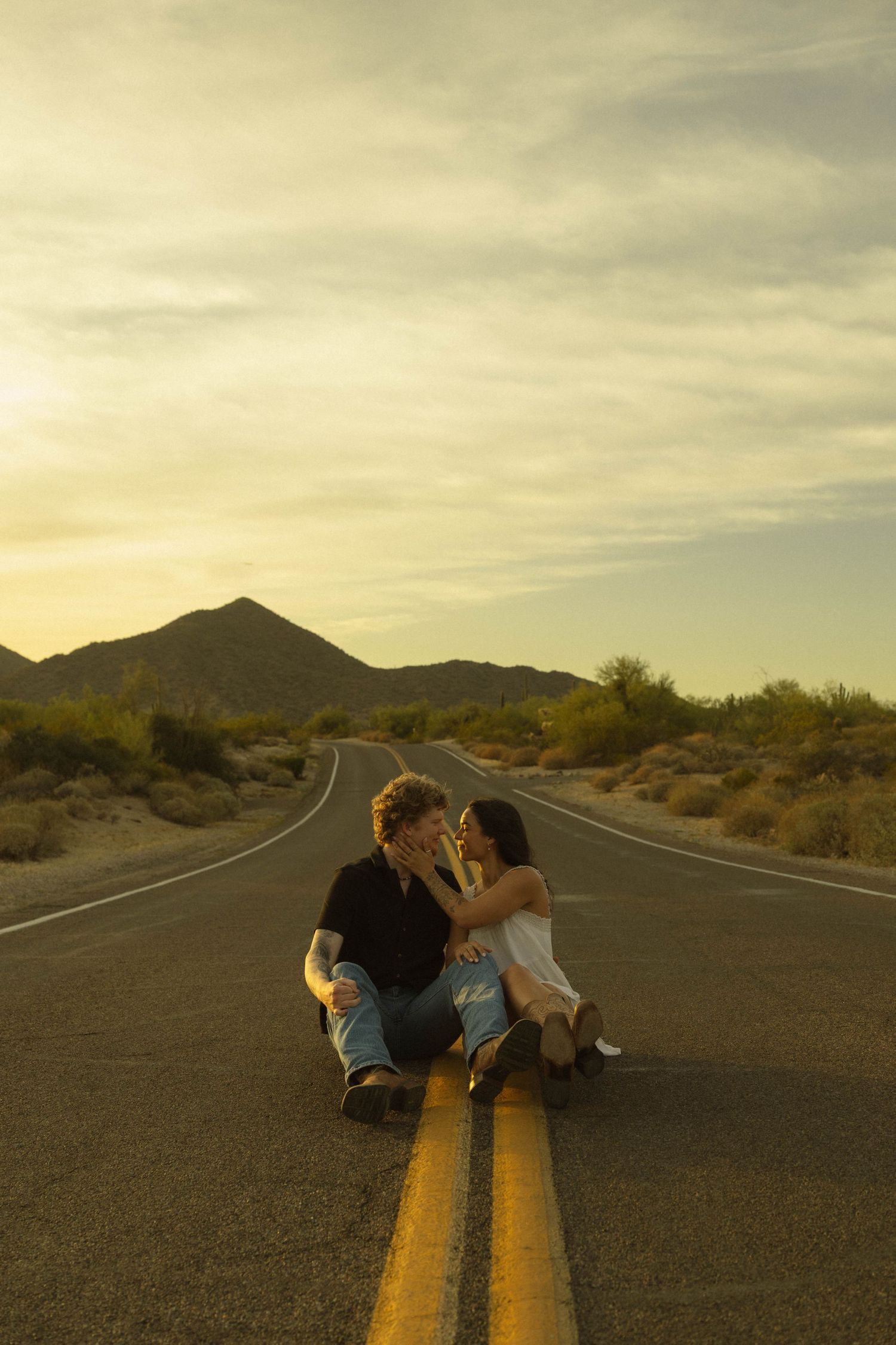 Couple sitting on desert road centerline at sunset with mountains in background.