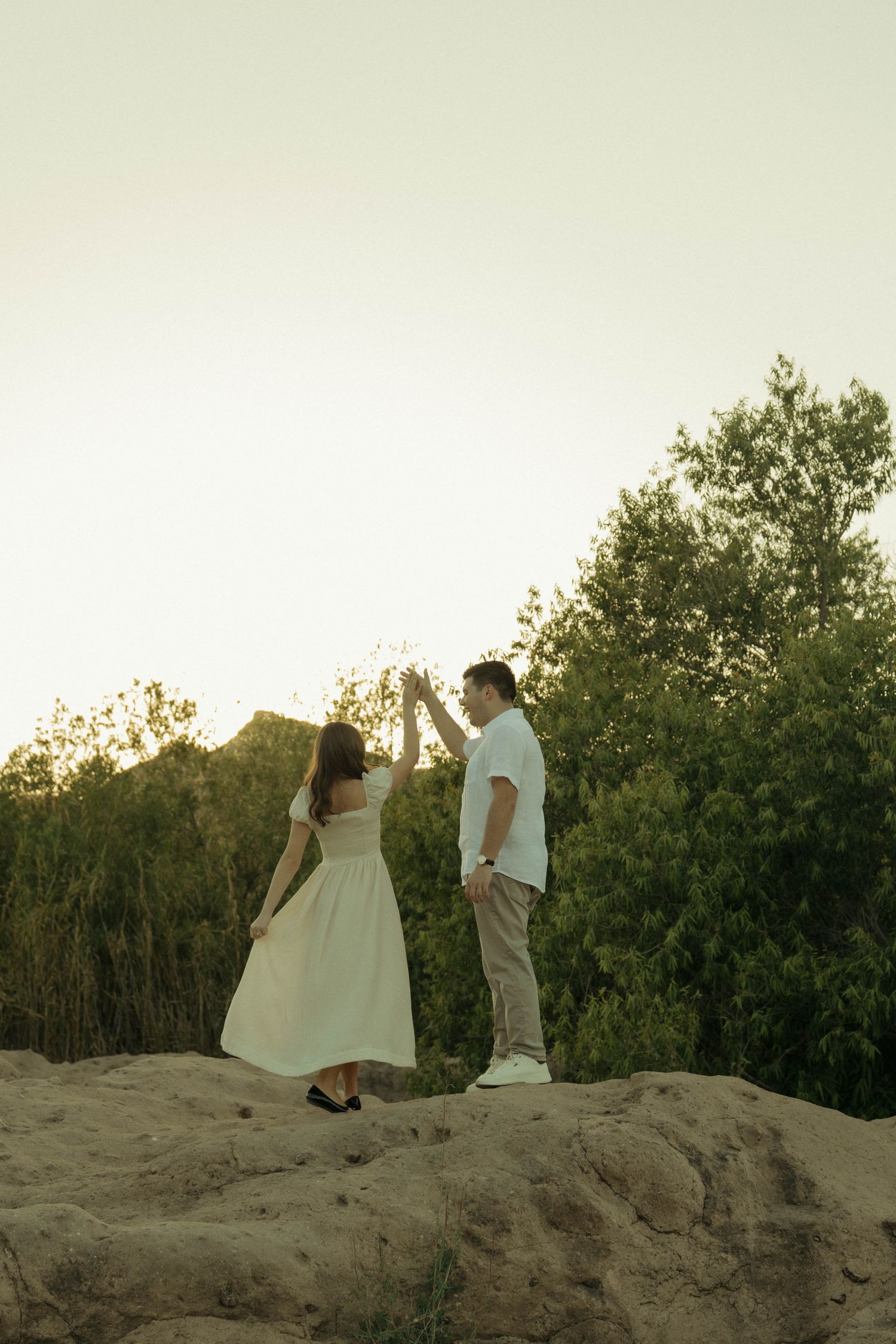 Couple dancing together on sandy hill at sunset with lush green trees in background.
