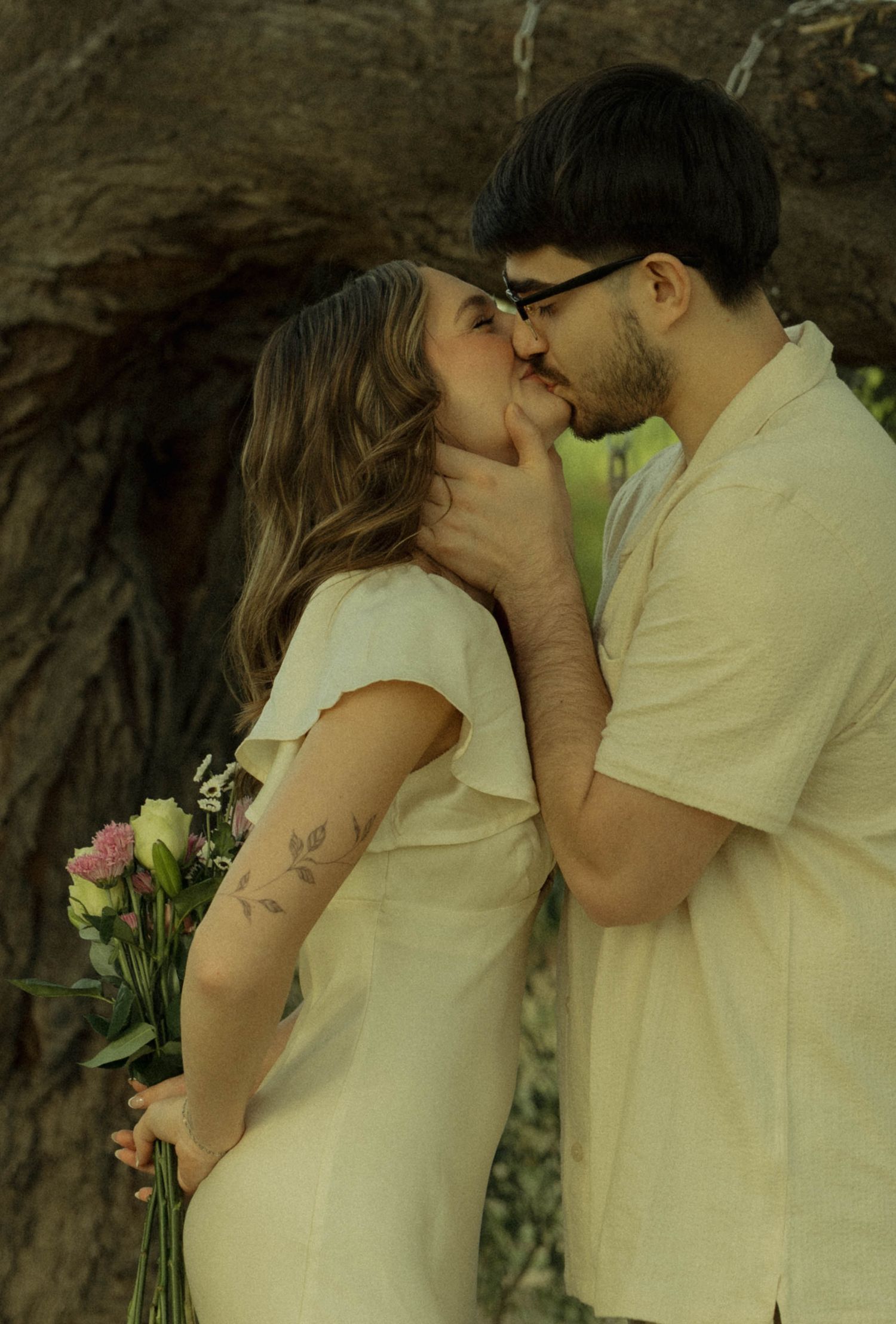 A couple shares a romantic kiss outdoors, the woman in a white dress holding a bouquet of flowers near a large tree.