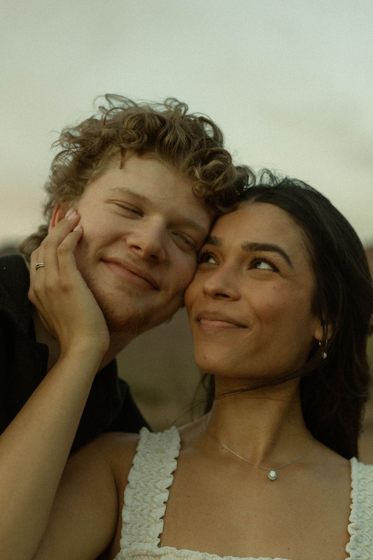Smiling couple in tender moment, woman in white top gently cupping mans face against soft sky backdrop.