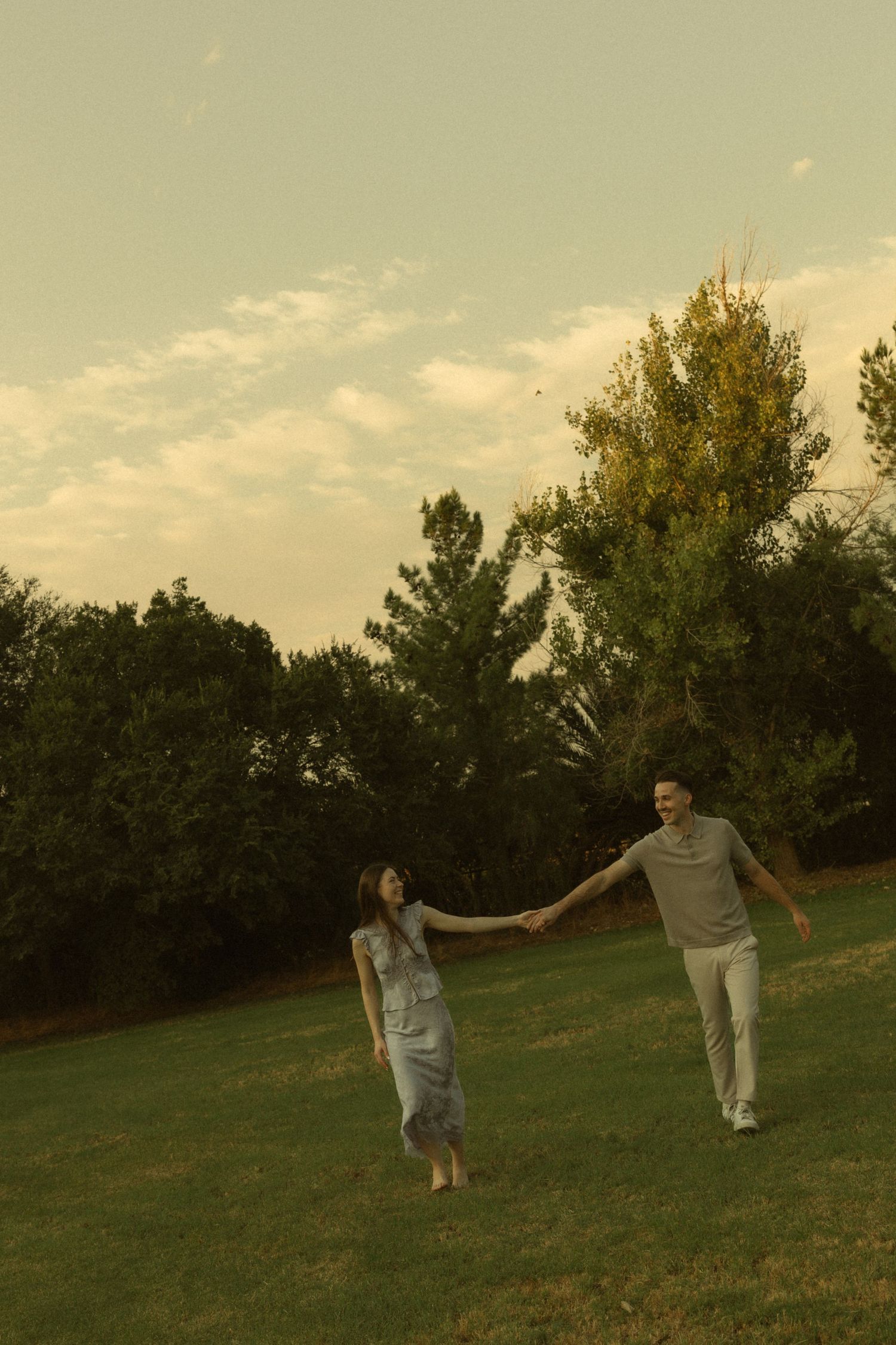 Couple holding hands walking on green grass field with trees and cloudy sky in background.