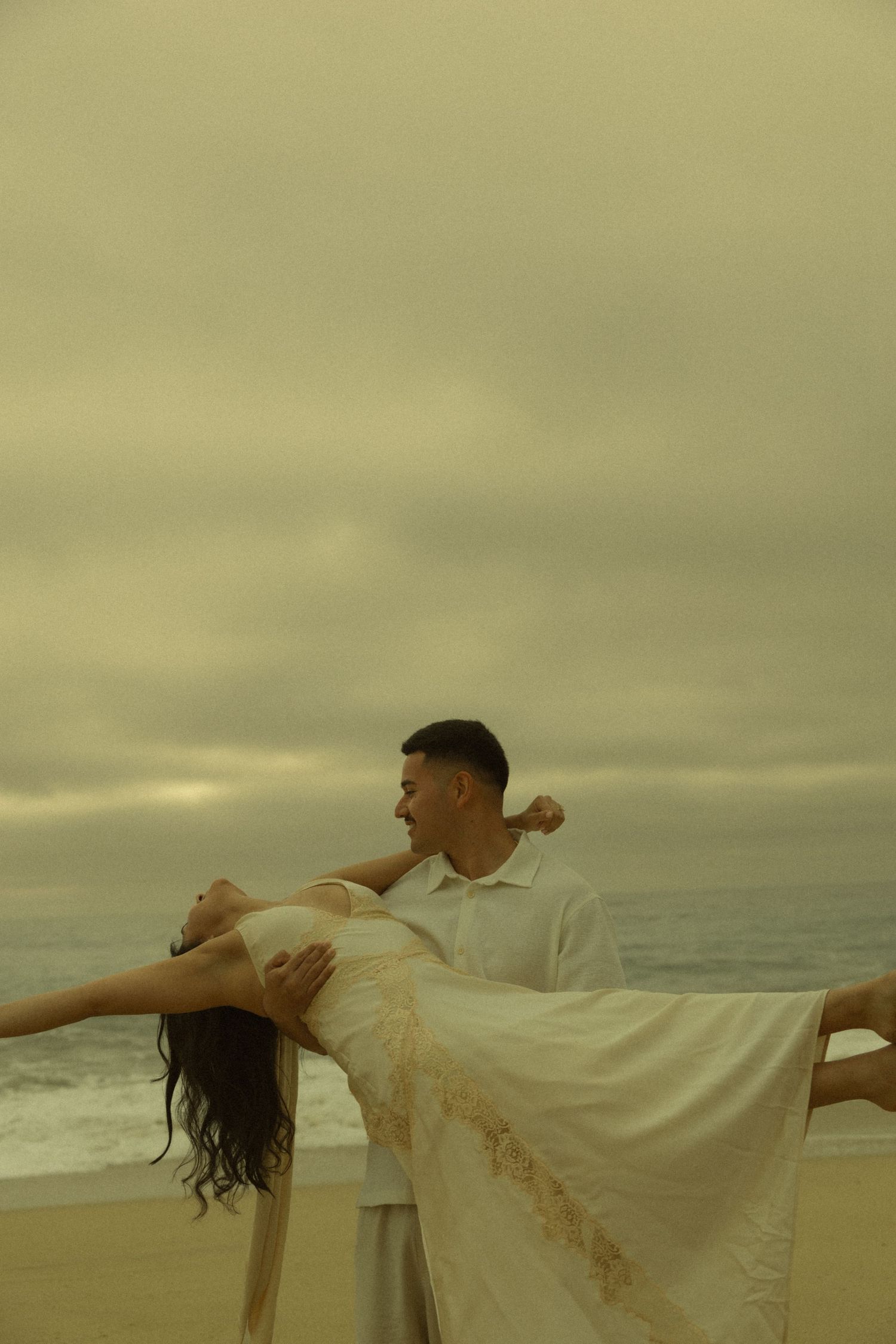 Man carrying woman in white dress on a cloudy beach with ocean waves in the background.