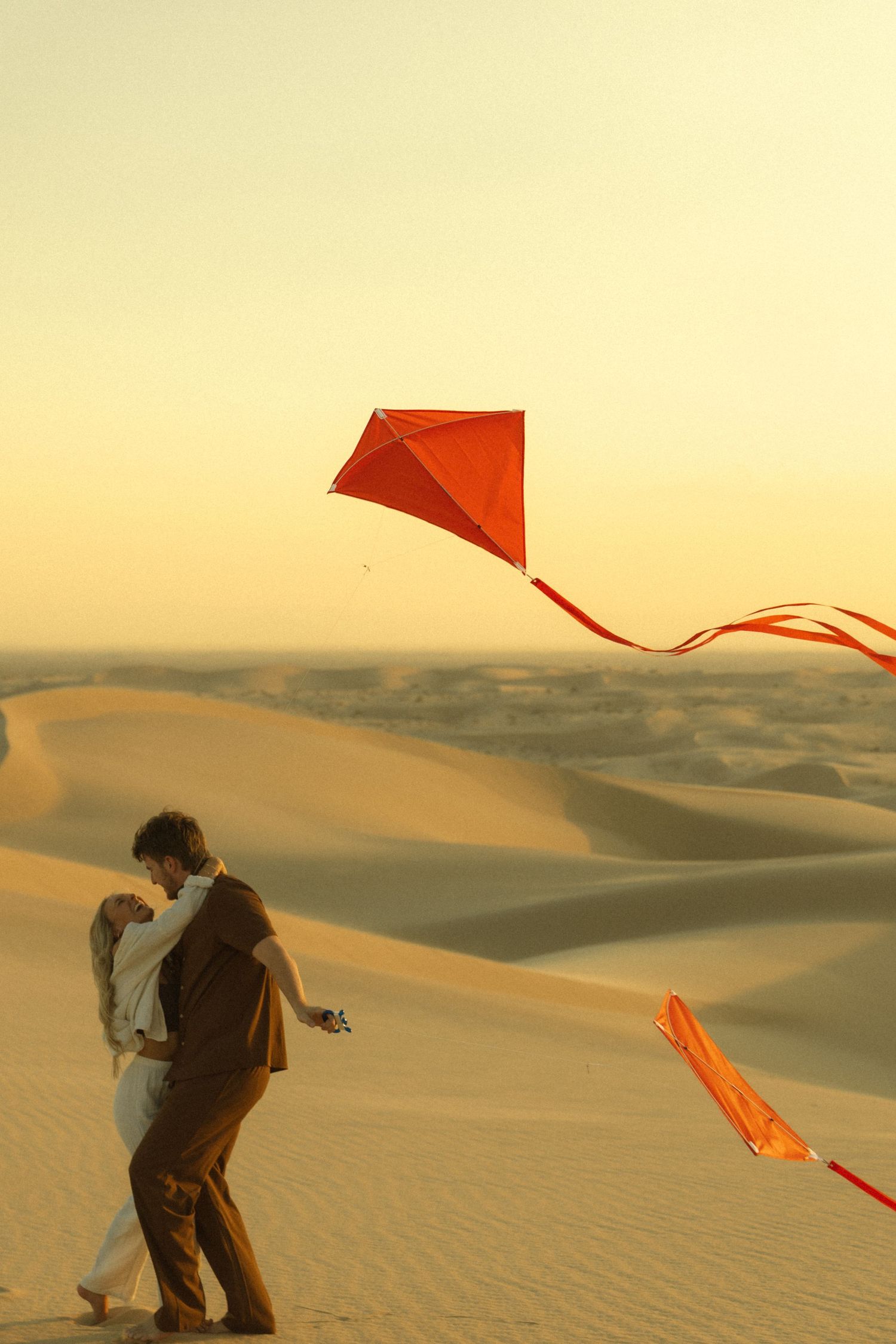 Couple embracing while flying a red kite on golden sand dunes at sunset in a vast desert landscape.