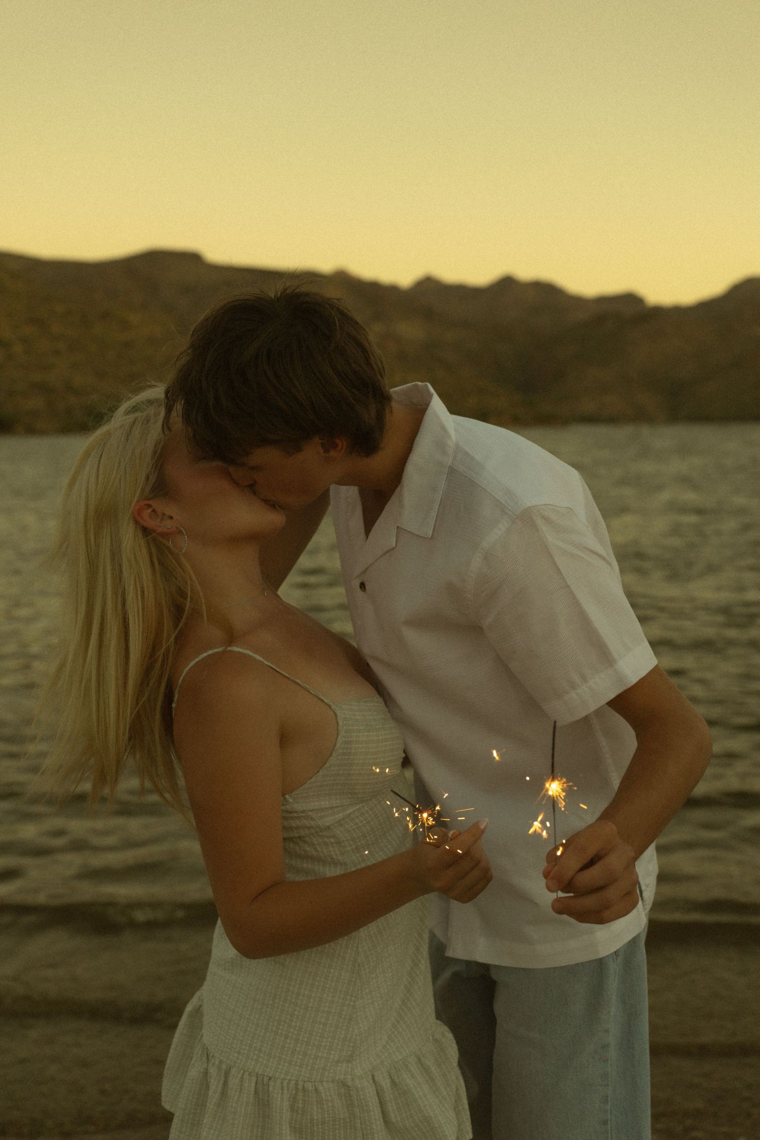 Couple kissing at sunset on a beach, holding sparklers, wearing white outfits with mountains in the background.