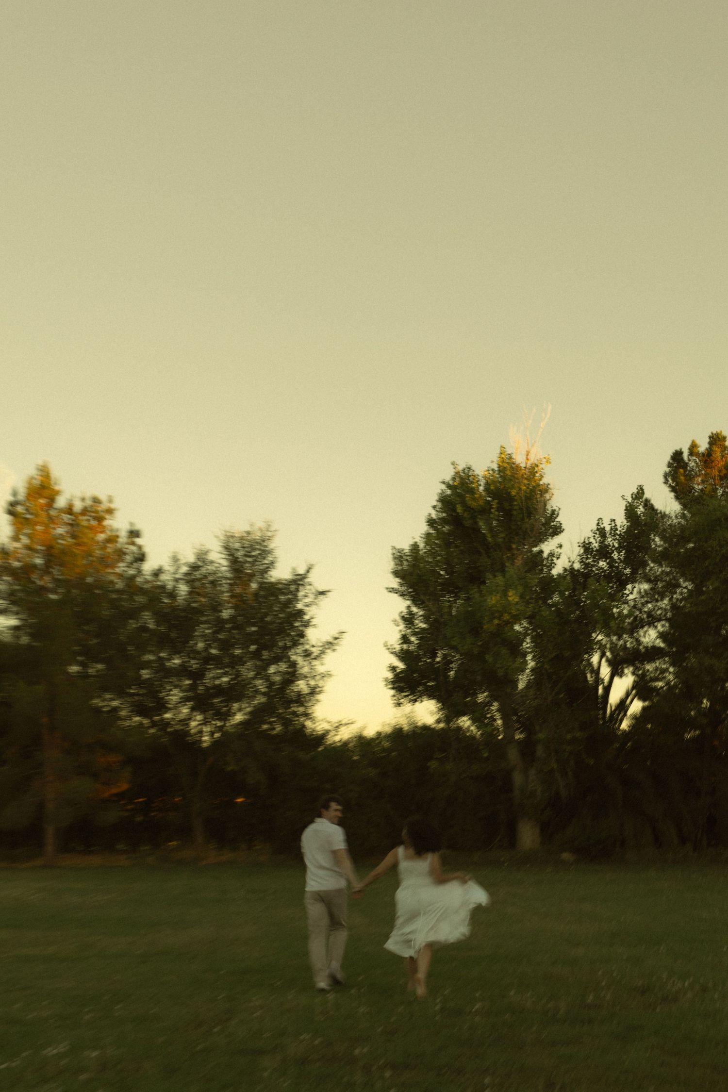 Couple holding hands running through a lush green park at dusk with tall trees in the background.