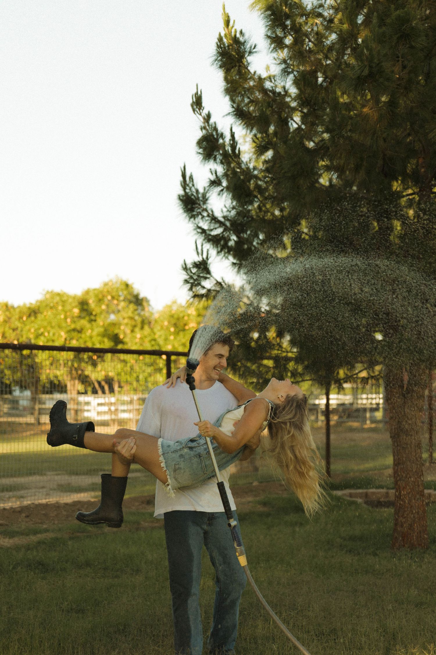A man carries a woman in his arms while holding a spraying garden hose in a sunny backyard.