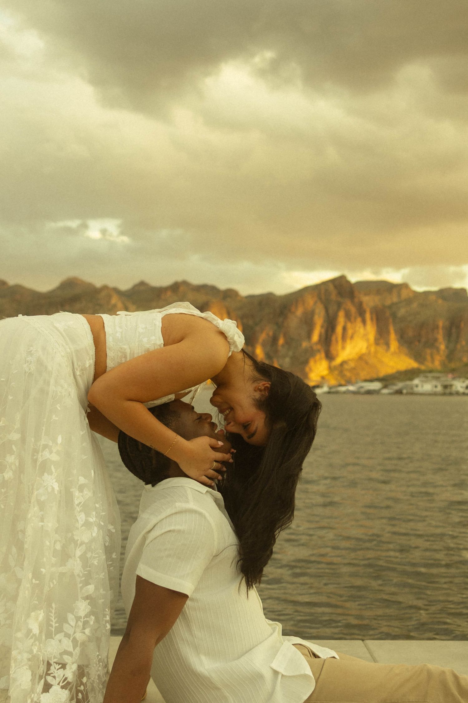Romantic couple sharing a tender moment by the water at golden sunset with dramatic mountain backdrop.