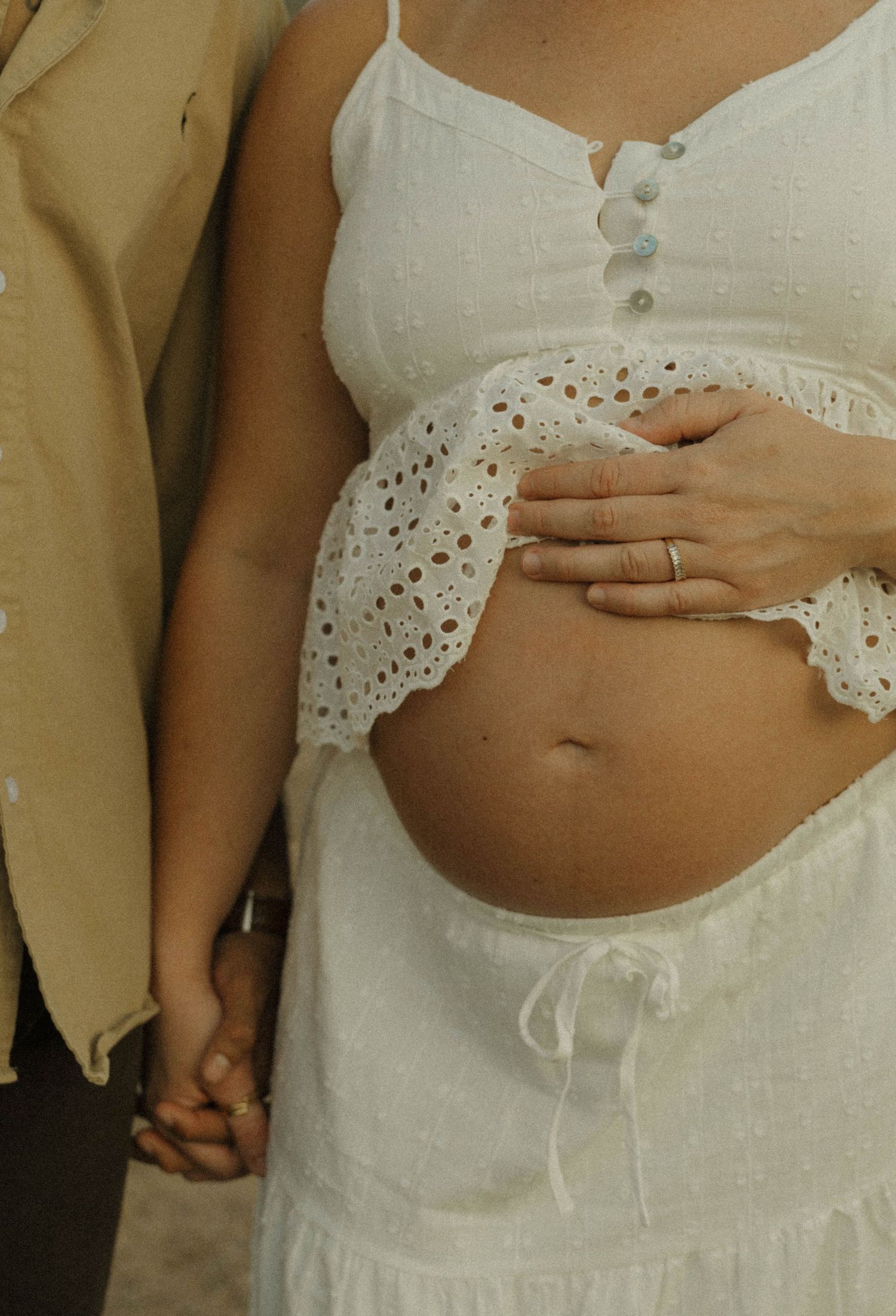 Pregnant woman in white eyelet dress holds baby bump while partner holds her hand outdoors.
