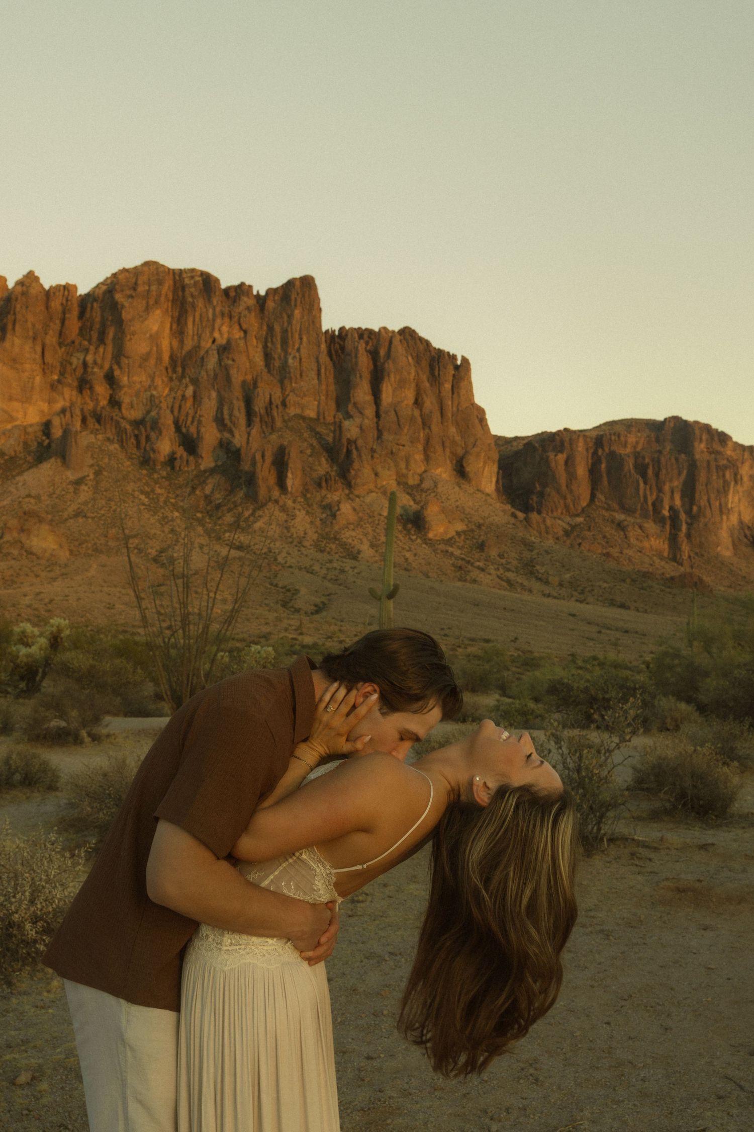 Couple sharing a romantic dip kiss at golden hour with Superstition Mountains in Arizona desert background.