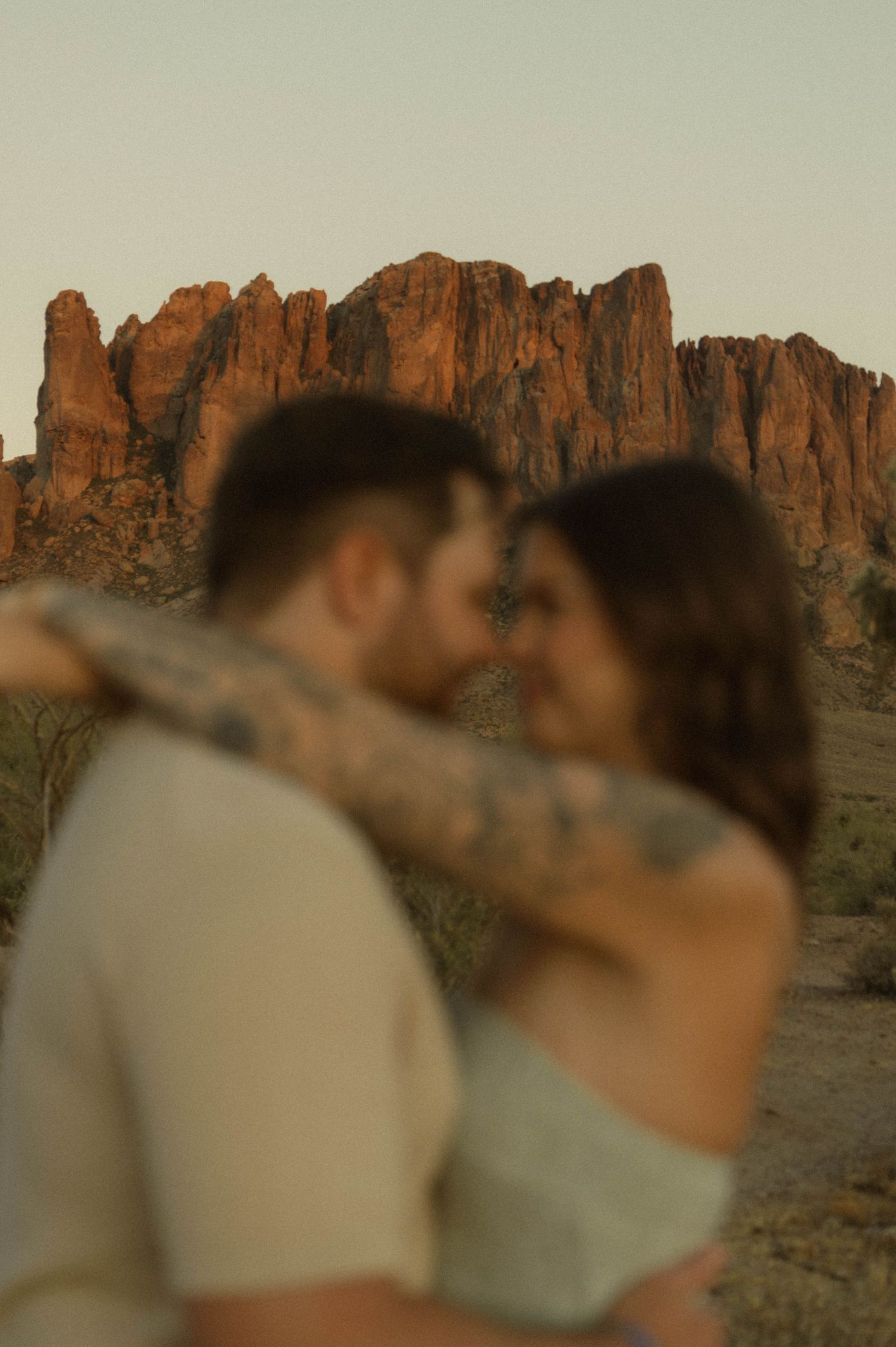 Blurred romantic couple embracing with tattooed arm visible against stunning red rock Superstition Mountains at sunset.
