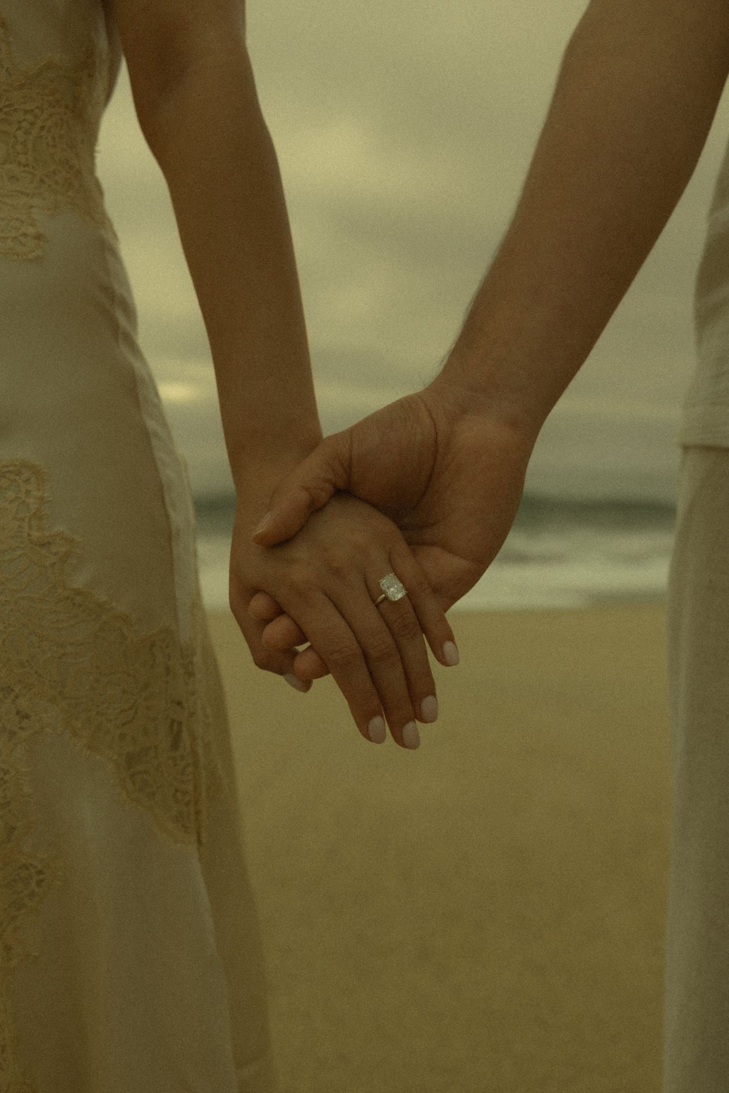 Close-up of couple holding hands on beach, woman wearing diamond engagement ring, overcast sky.