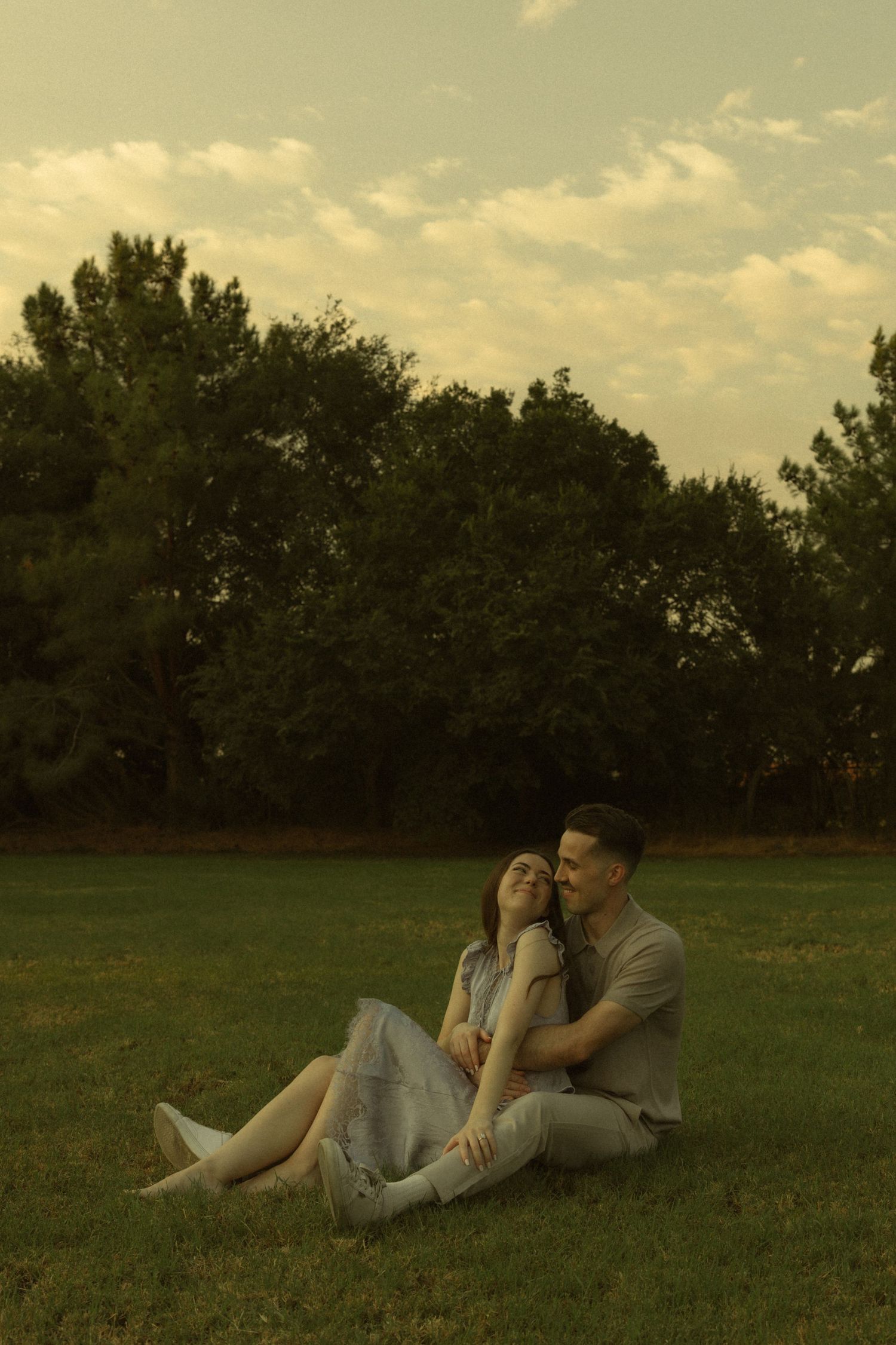 Couple sitting together on green grass at sunset, woman leaning on man with trees and cloudy sky behind them.