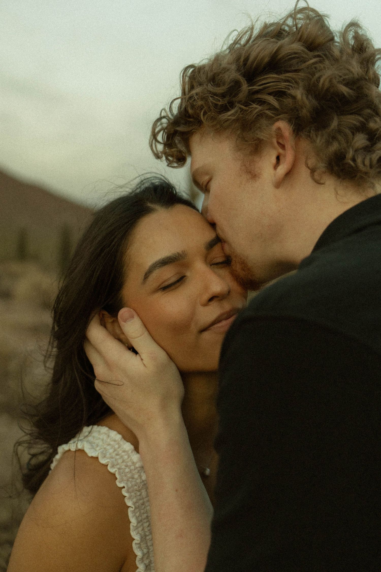 Curly-haired man tenderly kissing dark-haired woman on cheek in romantic desert engagement photo.