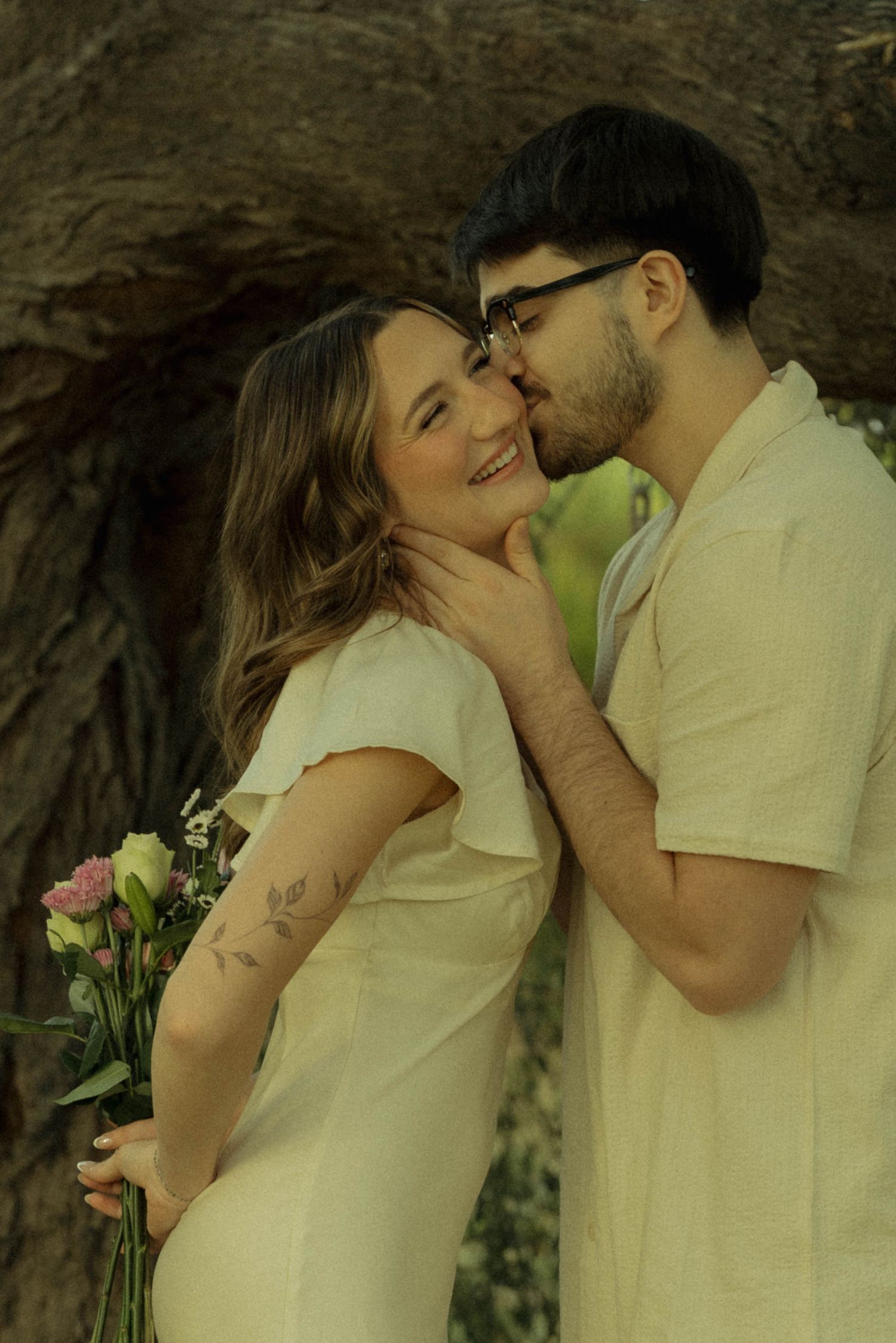A couple shares a romantic moment outdoors, man kissing woman's cheek as she smiles holding flowers near a tree.