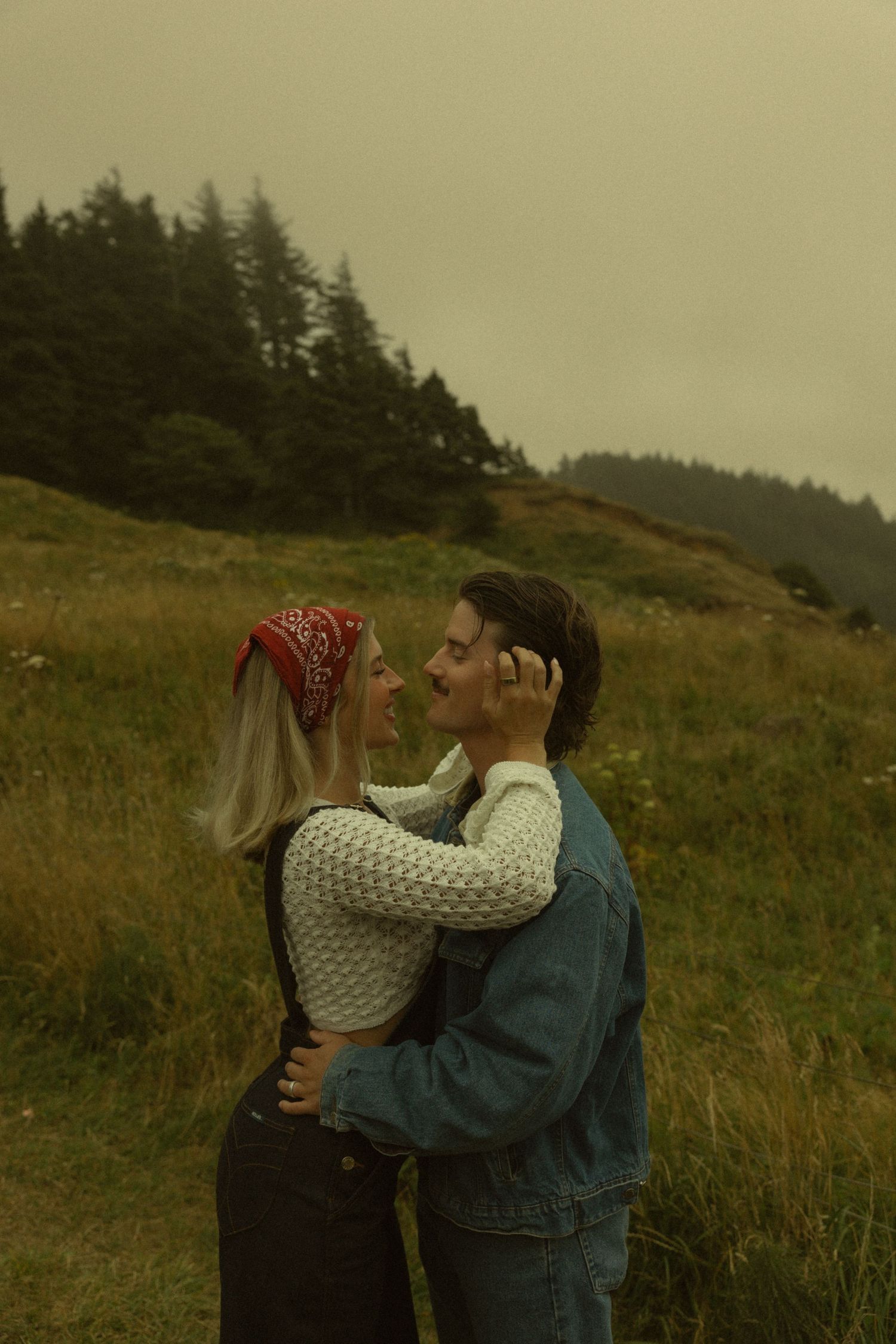 Couple embracing in a misty green meadow, woman in red bandana and white sweater, man in denim jacket, surrounded by pine trees.