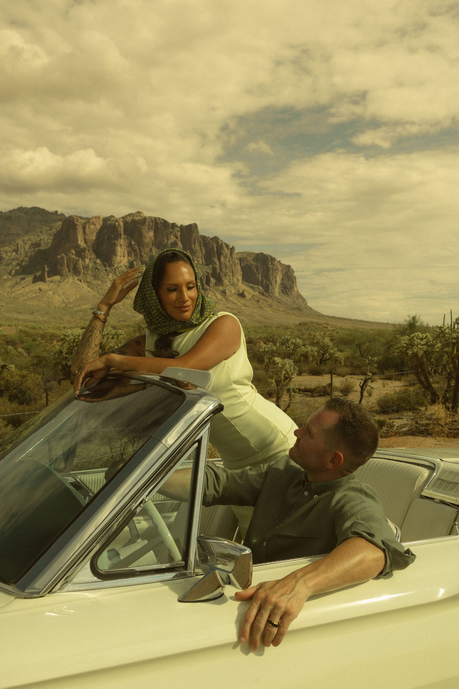 Couple in vintage convertible car with desert mountains backdrop, woman in green headscarf leaning on windshield.
