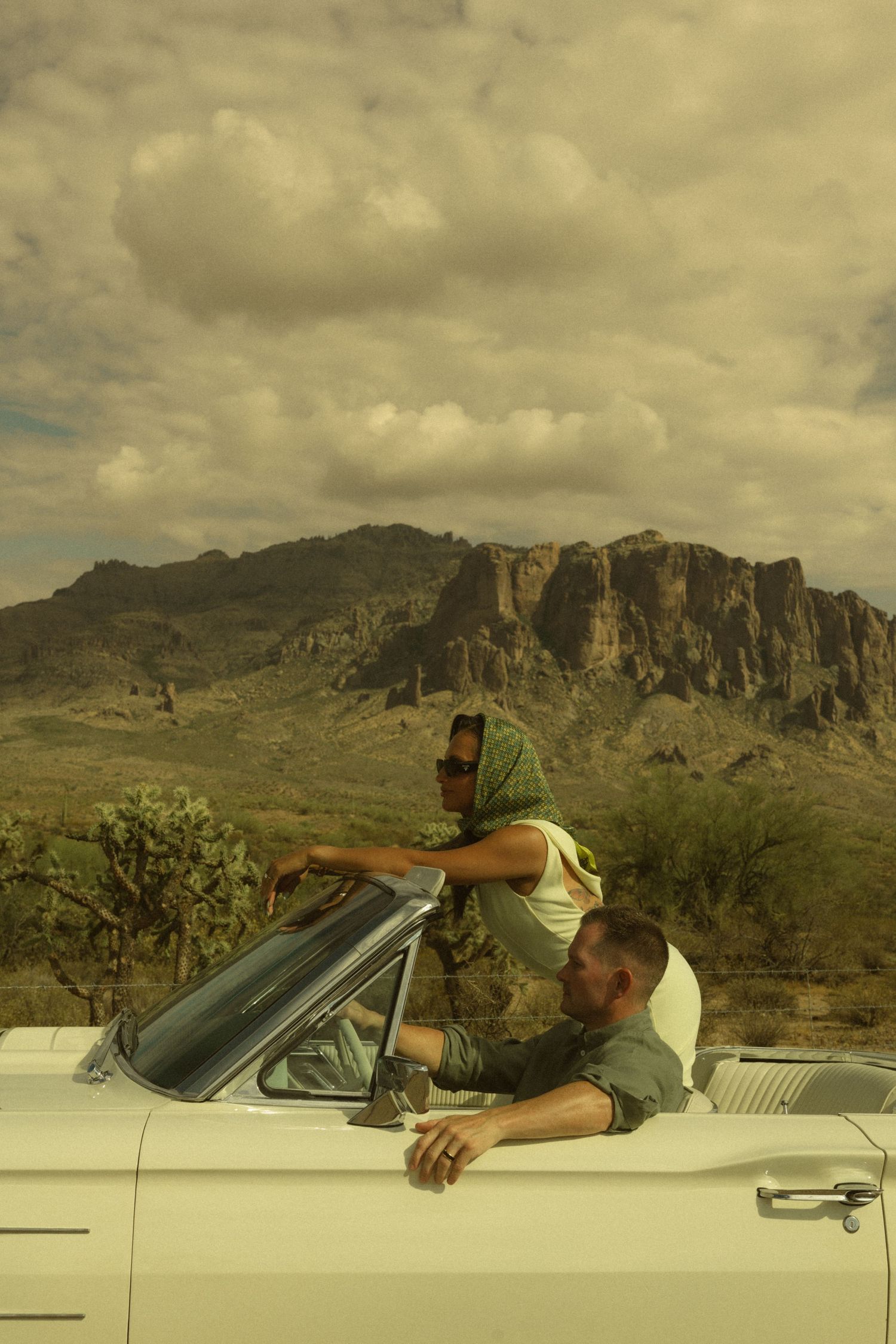Two people in a vintage white convertible car with dramatic desert mountains and cloudy sky backdrop.