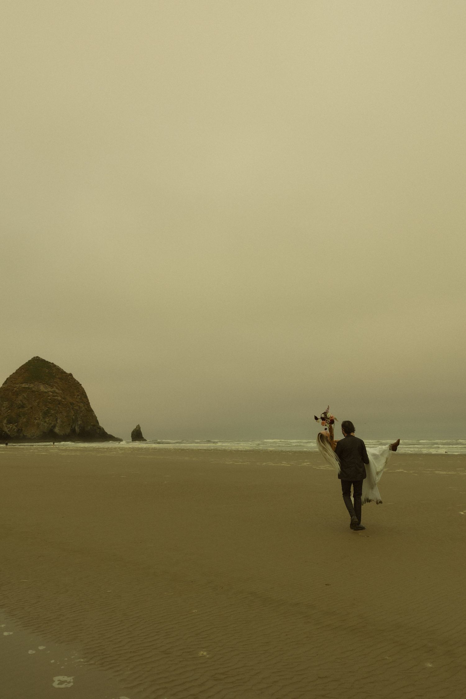 Groom carrying bride across sandy Cannon Beach with Haystack Rock in misty background.