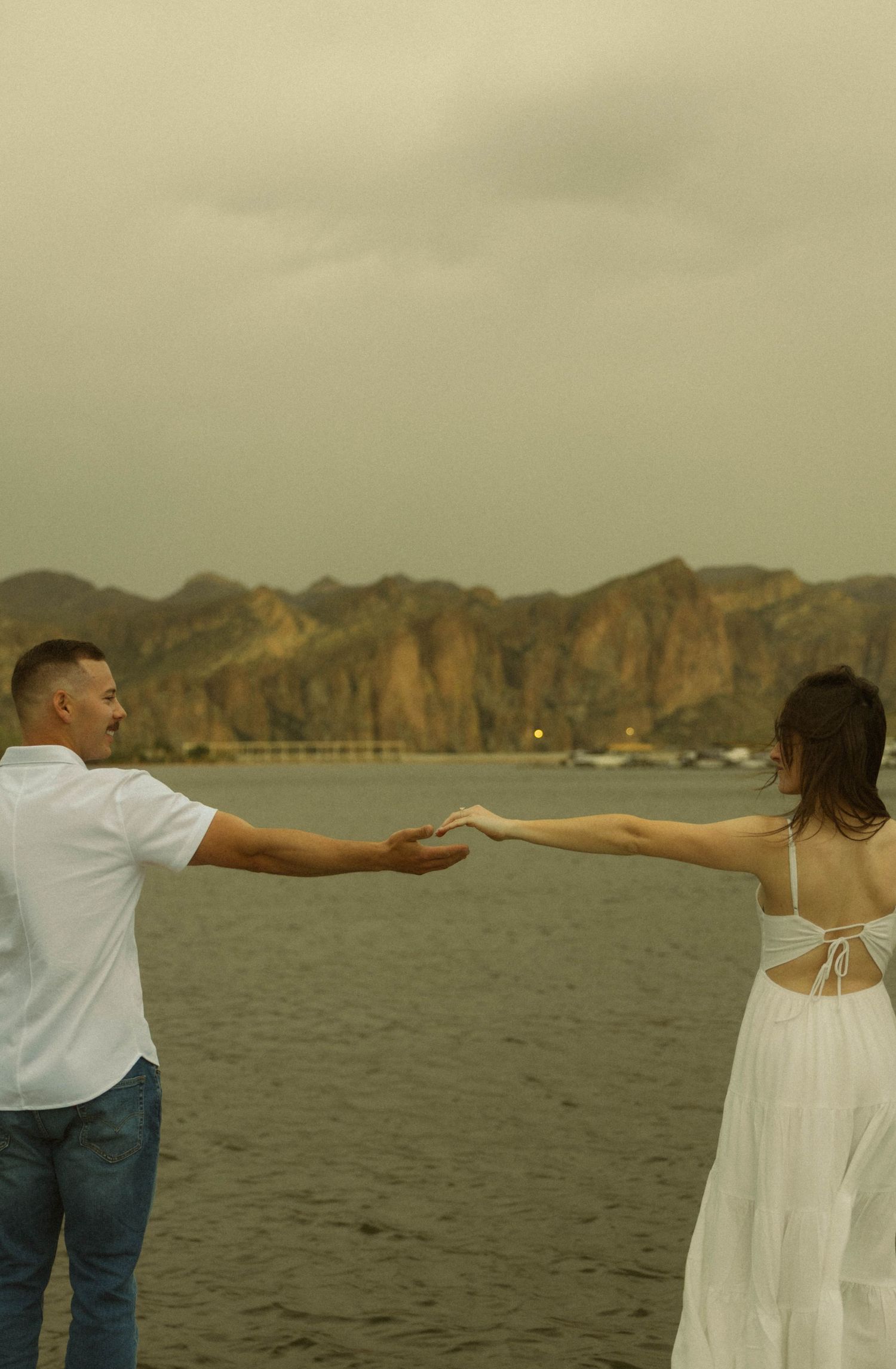 Couple holding hands while facing mountain vista near water.